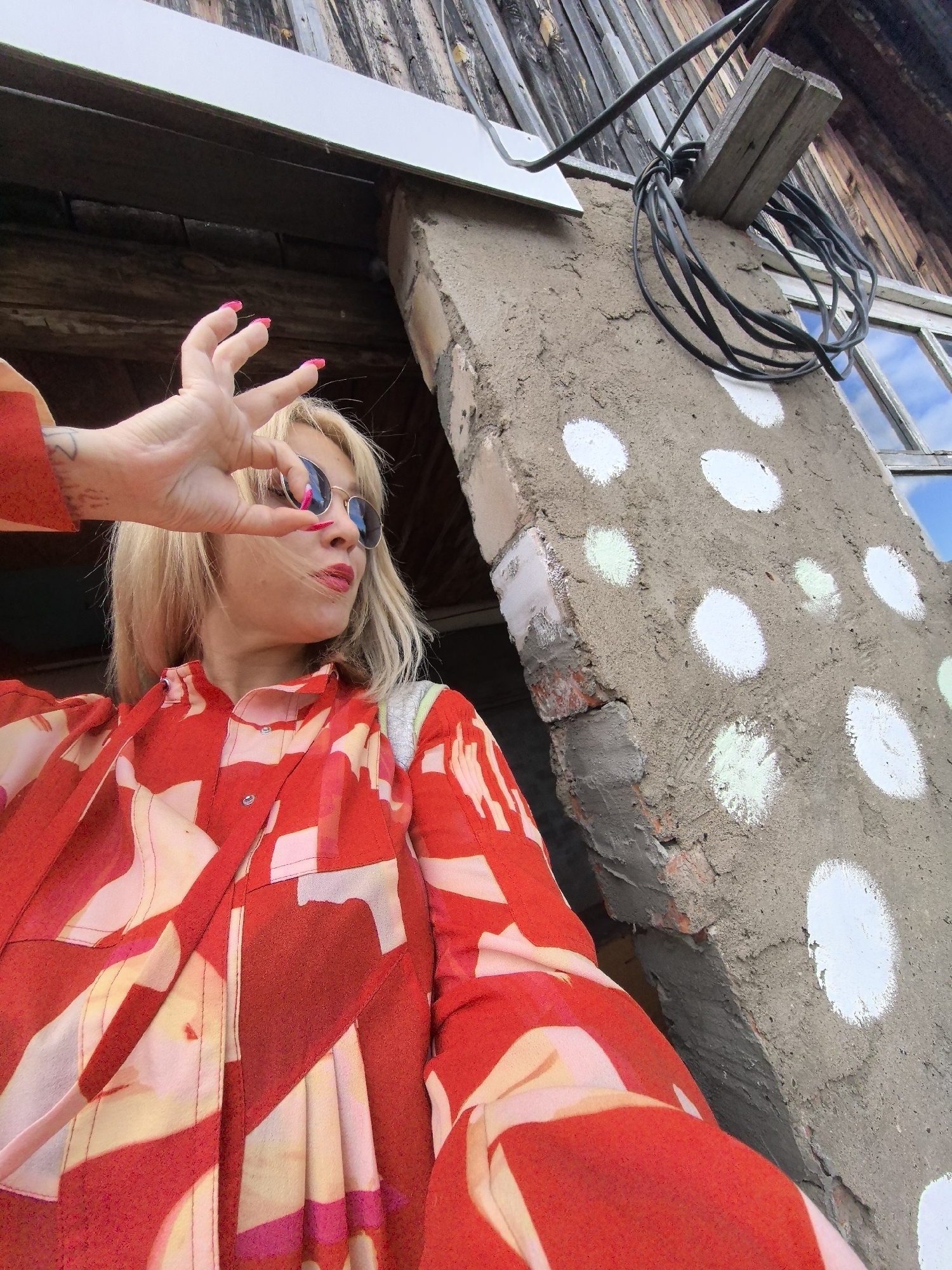 A blonde woman wearing sunglasses and a red and white patterned shirt poses with her hand in front of her face, obscuring her eyes. She is standing outside a building with a weathered wooden exterior and a concrete wall decorated with white and green polka dots. The building appears to be in a rural or semi-rural location, with exposed brick and construction materials visible. A coiled black cable is attached to the concrete wall. The scene suggests an informal, candid moment, possibly taken in Noviy, Russia, given the context.