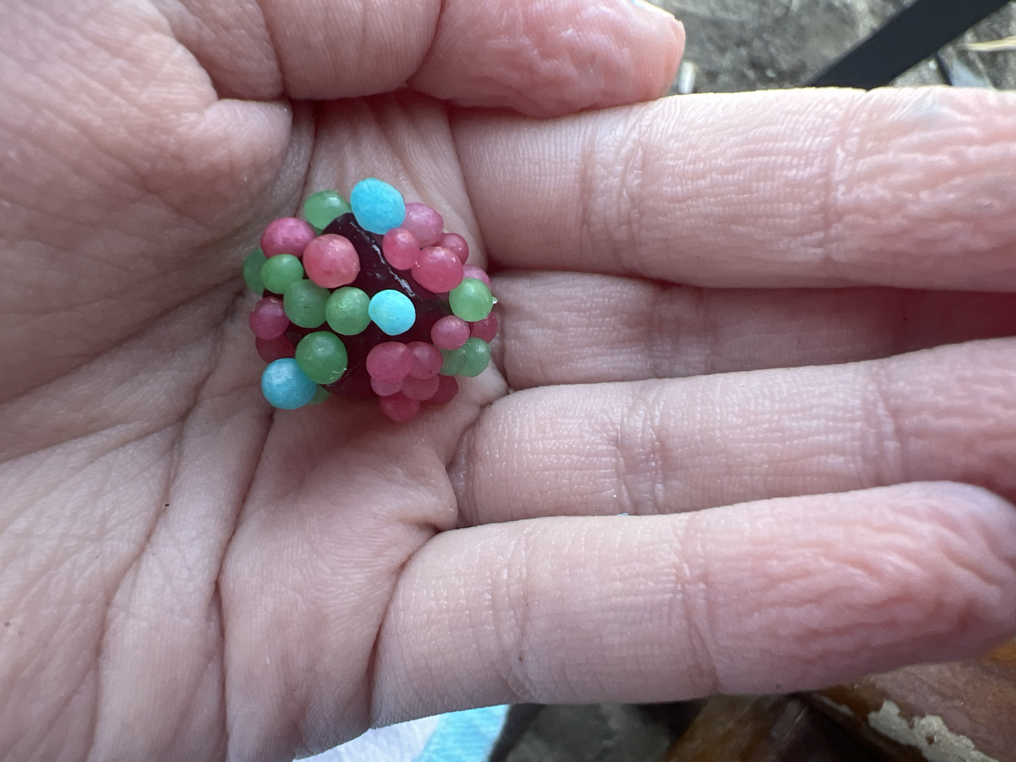 The image shows a close-up view of a person's palm holding a cluster of small, colorful candies. The candies are round and appear to be of various colors, including pink, green, and blue, with a dark purple base. The palm is lightly wrinkled, indicating the texture of the skin. The background is out of focus but suggests an outdoor environment with some brown and gray elements, possibly wood or stone. There is no discernible urban environment visible, nor any specific landmarks that would indicate the location within Eretria Municipal Unit, Greece.