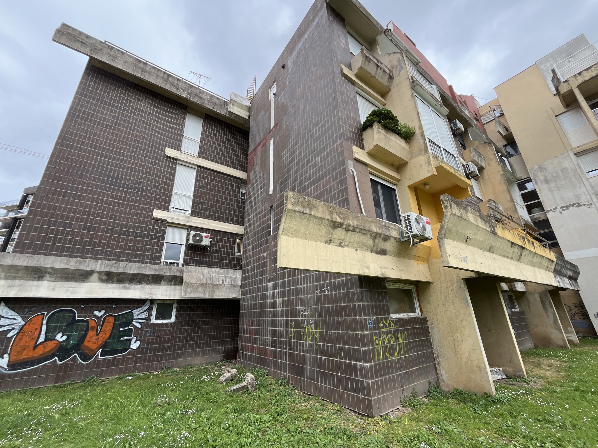 The image shows an exterior view of a multi-story building in Budva, Montenegro. The building has a mix of brown tiled sections and weathered yellow painted concrete. Graffiti, including the word "LOVE" with winged hearts, is visible on the lower part of the brown tiled wall. The ground is covered in grass and small white flowers. Several air conditioning units are mounted on the exterior walls. The sky is overcast.