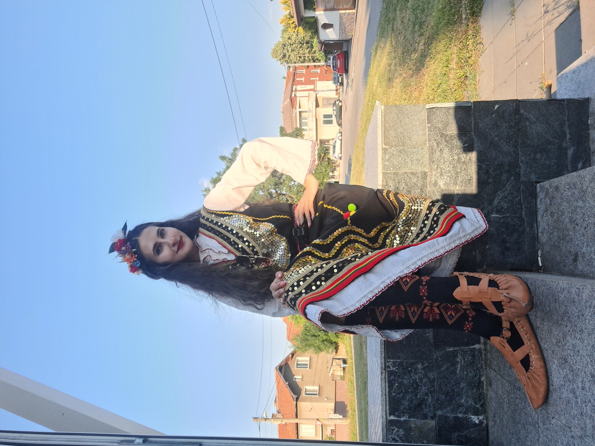 A young woman with long, dark, wavy hair and a floral headband is sitting on a stone staircase outdoors. She is wearing a white blouse with red embroidery and a black vest adorned with gold sequins and colorful pom-poms. Her skirt has a white hem with red and gold trim, and she is wearing black patterned leggings and brown leather shoes. The background shows a street with houses, trees, and cars under a clear blue sky.
