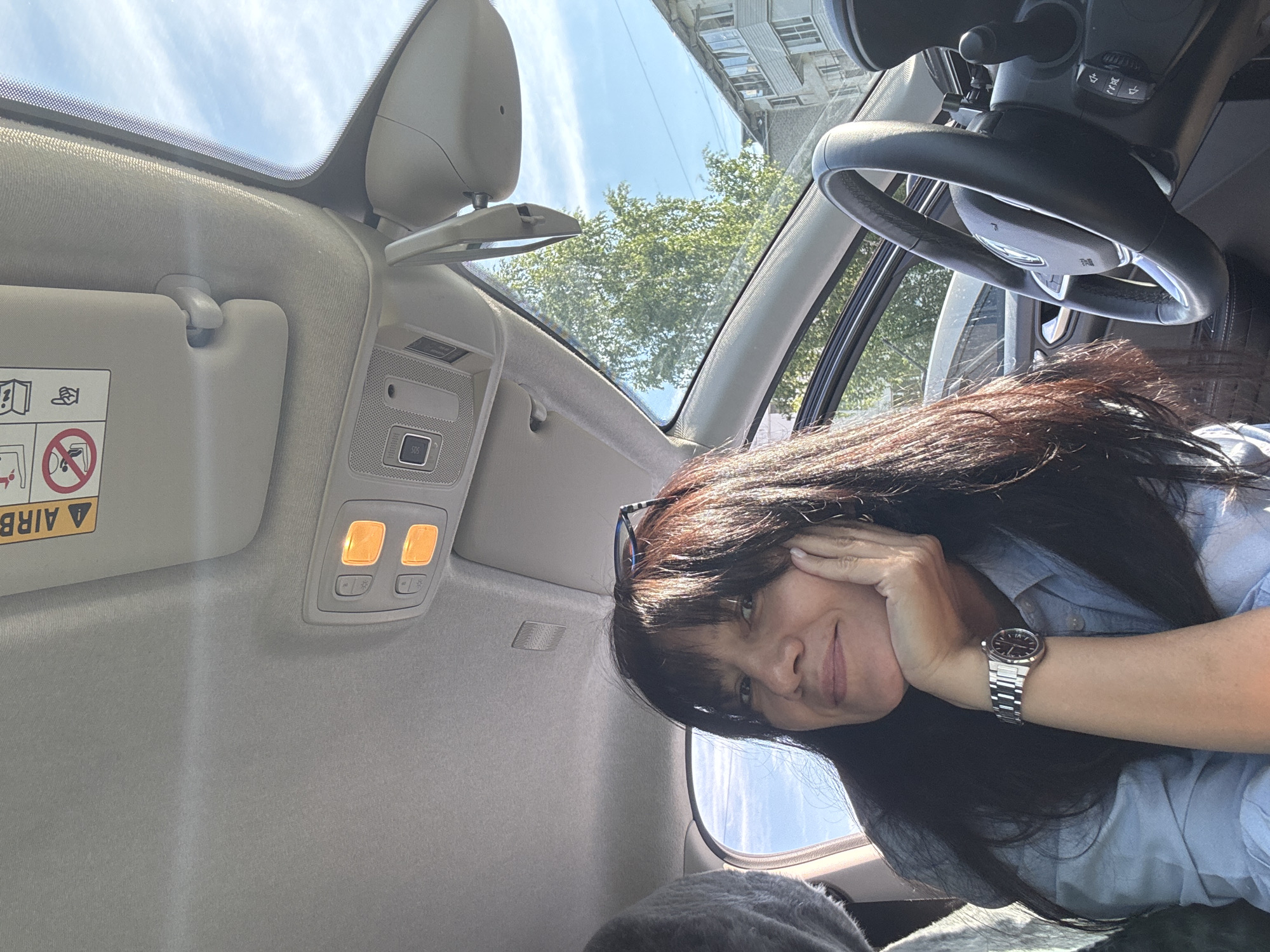 A woman with long, dark hair and sunglasses perched on her head is captured inside a car during what appears to be a sunny day in Chaikovsky, Russia. She is shown from a high angle, looking towards the camera with a slight, relaxed smile, resting her head on her right hand. She wears a light blue button-down shirt and a silver-toned watch with a metal bracelet on her left wrist.
The car's interior features a light-colored headliner and a sun visor with upside-down warning labels, including the word "AIRBAG" and various safety symbols. An overhead console is visible, illuminating two amber lights and featuring several buttons, notably one marked "SOS." Through the car's large windshield or panoramic roof, a clear blue sky with a few clouds is seen, along with bright green trees and the upper floors of a building, likely residential. A portion of the car's dark steering wheel is visible on the right, showing some controls. The scene conveys a calm, bright daytime atmosphere.