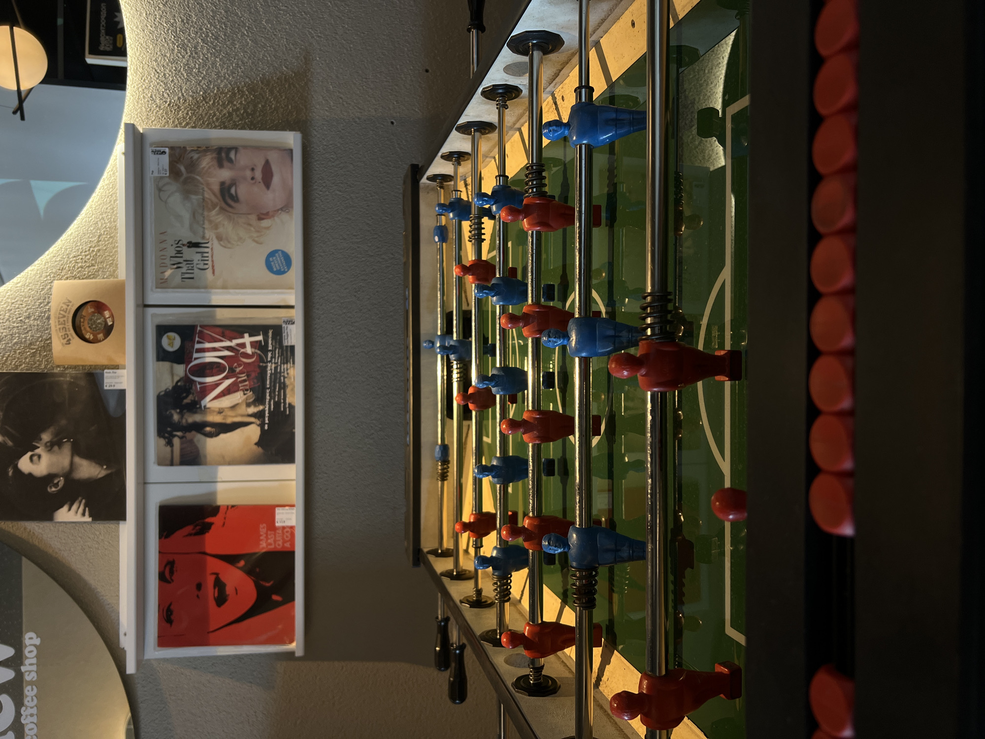 The image showcases a detailed view of a foosball table, positioned near a wall adorned with record sleeves. The foosball table has the classic red and blue players mounted on silver rods, with a green playing field visible in the background. The wall features a collection of records, including titles like "Who's That Girl" by Madonna and an album by James Last. A portion of a sign or structure with the text "coffee shop" can be seen on the left side of the frame. The setting is likely an indoor space, possibly in a commercial establishment. The location is not explicitly stated in the image; "Rafailovići, Montenegro" is external information.