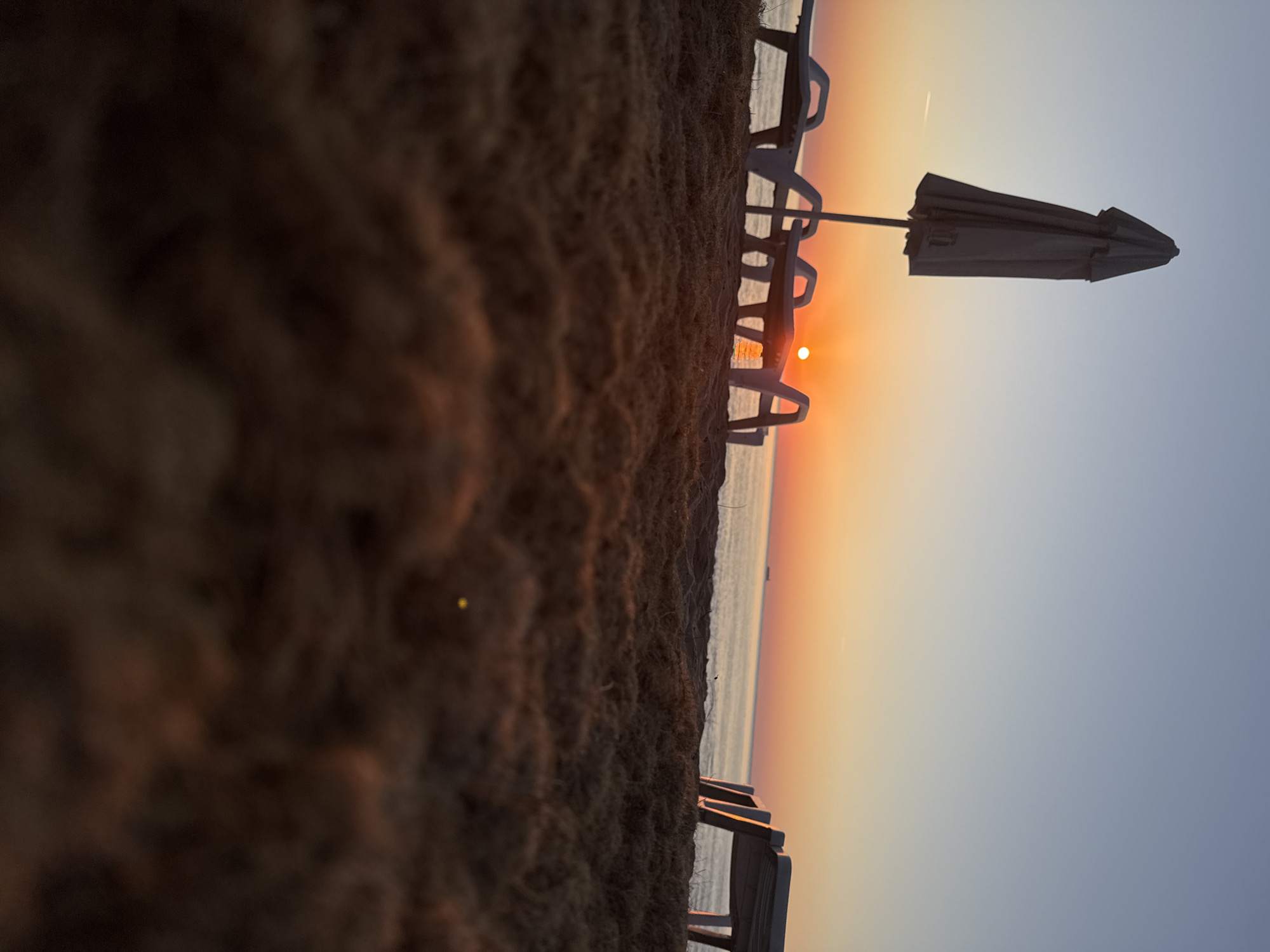 The image captures a serene beach scene at sunrise in Shkorpilovtsi, Bulgaria. In the foreground, the textured sand is out of focus, creating a shallow depth of field. Beyond the sand, several white plastic beach chairs and a closed beach umbrella are arranged on the sand. The sun, a bright orange orb, is visible just above the horizon, casting a warm glow over the sky and the calm sea. The sky transitions from a pale blue at the top to shades of orange and pink near the horizon. A faint contrail from an aircraft can be seen in the upper left portion of the sky. A distant ship is visible on the horizon in the upper right of the image. The overall atmosphere is tranquil and suggestive of the early morning hours at a seaside resort.