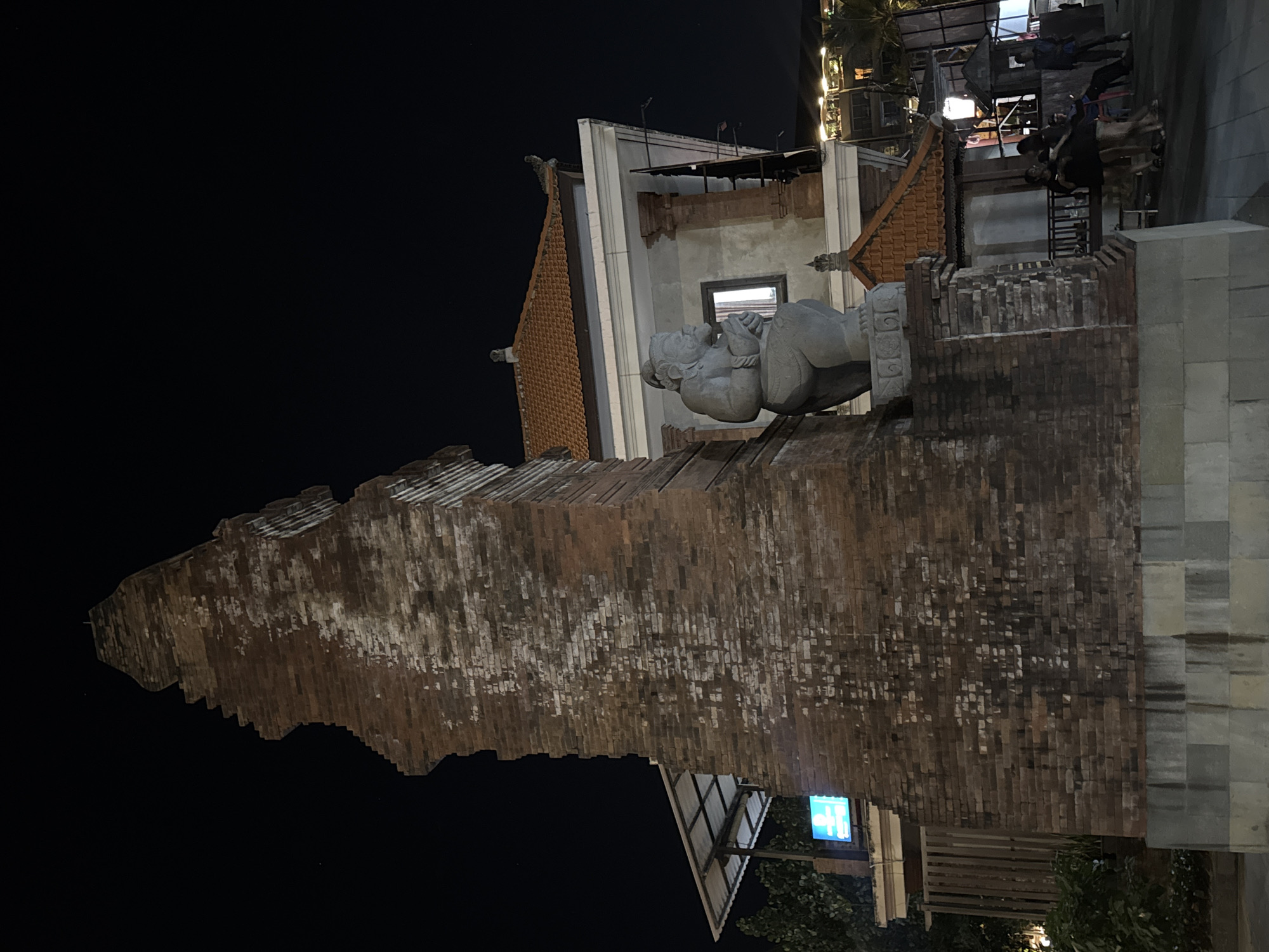 This is a nighttime photograph of a large, intricately carved brick structure in Kuta, Indonesia. The structure appears to be an entryway or part of a building, with a traditional Indonesian architectural style characterized by a steep, tiled roof and decorative carvings.
Dominating the foreground is a stone statue of a seated figure, possibly a deity or mythical being, depicted with its hands clasped together. The statue is positioned atop the brickwork, contributing to the imposing presence of the structure. The brickwork itself is weathered and shows signs of age, with patches of white and darker tones interspersed among the reddish-brown bricks. The top of the brick structure has a jagged, uneven edge.
Behind the brick structure, a building with white walls and orange tiled roofs is visible. A window on this building is illuminated, suggesting it is occupied. In the background, more buildings and lights are visible, indicating a developed area, likely a street or plaza. There are also several people in the background, some appearing to be walking or gathered.
The image captures a scene at night, with the darkness of the sky contrasting with the illuminated areas of the buildings and streetlights. The overall mood is atmospheric and hints at the cultural and architectural richness of the location. A blue sign with white text is partially visible on the lower right side of the image.