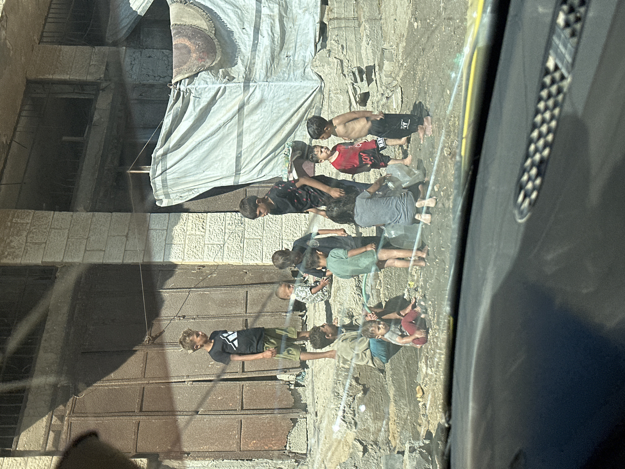 A group of children are gathered on a concrete ledge in front of a weathered metal door. One boy, wearing a black t-shirt and shorts, stands on the upper ledge. Several other children are on a lower, rougher concrete area, some sitting, some standing. One boy in a red shirt and black shorts is talking to another bare-chested boy in black pants. A younger child in a grey shirt and red pants sits on the ground. A green hose is visible, suggesting water is being used, perhaps for play. The background shows a brick building with a large, draped tarp. The scene is outdoors, in what appears to be a modest urban environment.