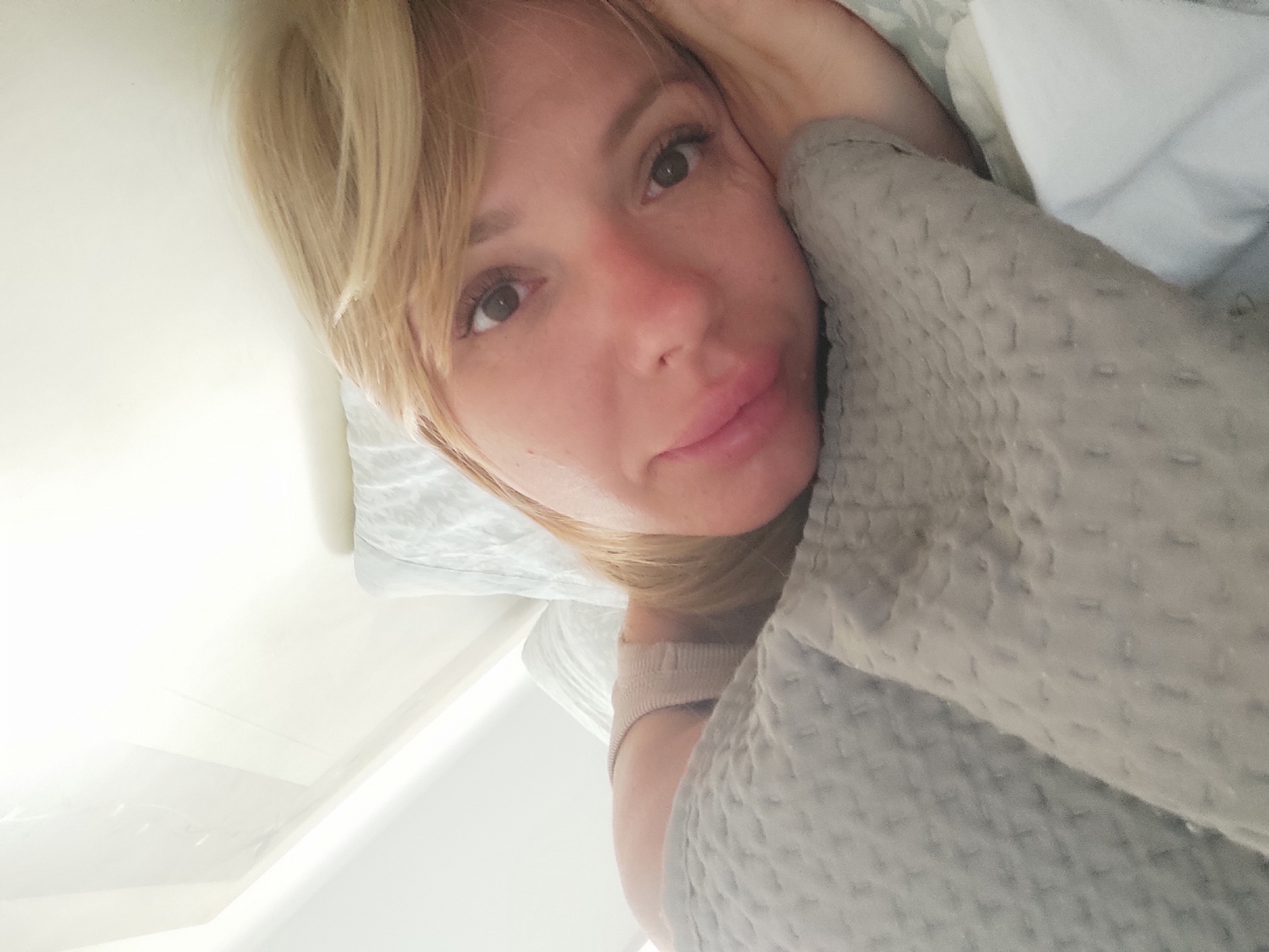 The image shows a close-up, upside-down, selfie-style shot of a person's face and upper body as they lie in bed. The person has blonde hair, is wearing a light-colored, ribbed sleeve that extends to the shoulder, and is partially covered by a textured gray blanket. The background suggests an indoor setting with a white wall and a light-colored pillow visible. The lighting appears to be natural and bright, possibly from a window. There is no discernible information in the image to suggest a specific location within Blanes, Spain.