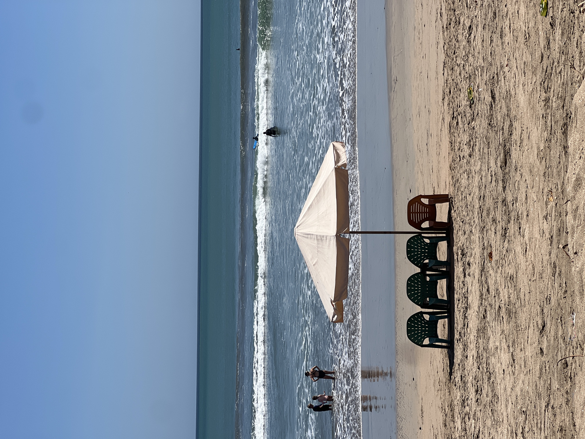On a sandy beach in Kuta, Indonesia, three green plastic chairs and one brown plastic chair are lined up behind a large, beige beach umbrella. The ocean, with gentle waves and a clear blue sky, is visible in the background. Several people are in the water: two children and an adult are standing near the shore, while further out, a surfer on a blue surfboard rides a wave, and another individual is wading. The beach is scattered with debris, including leaves and plastic.
