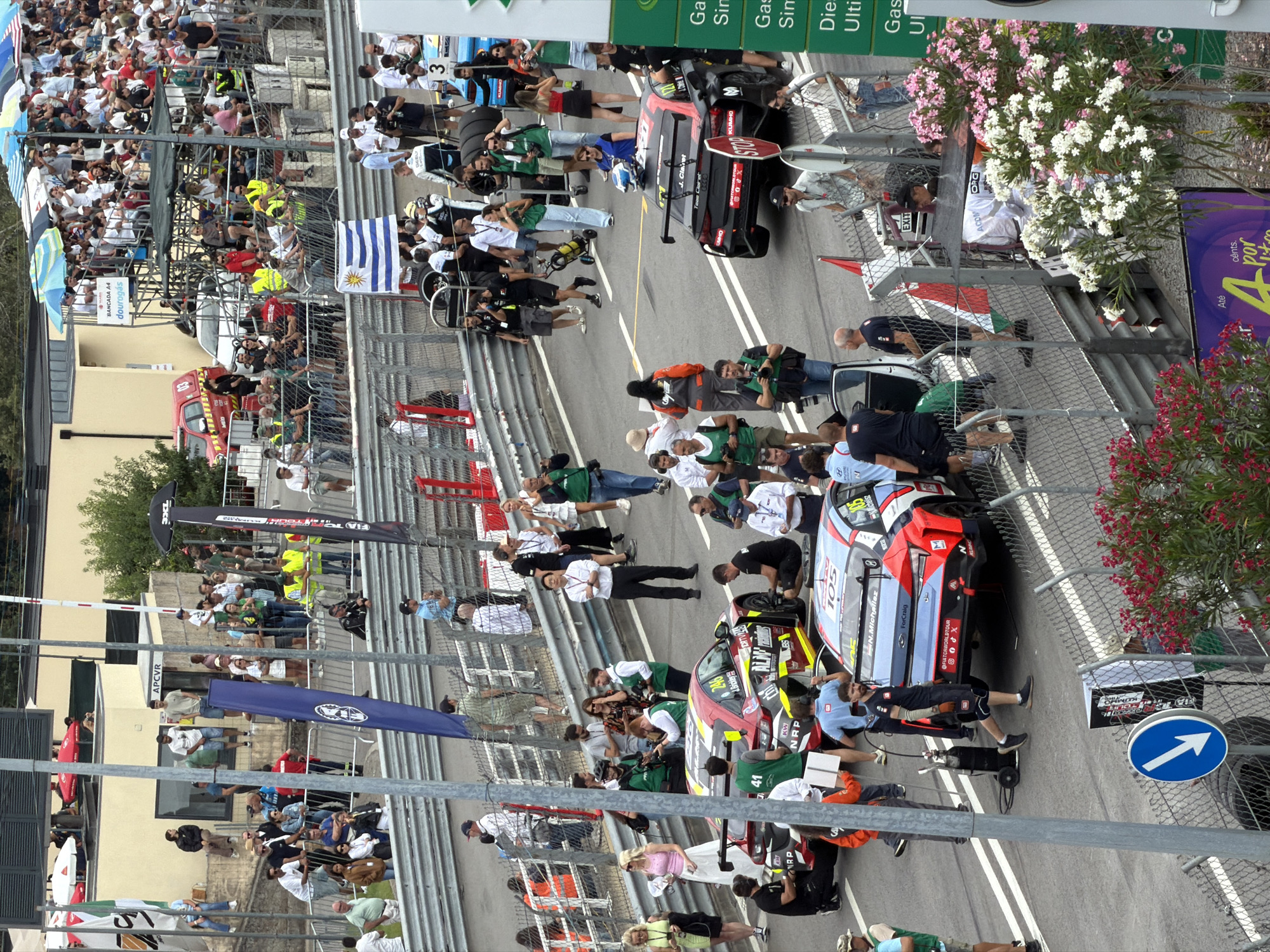The image captures a scene at the Circuito de Vila Real, a street circuit in Vila Real, Portugal, during a racing event. In the foreground, several race cars are parked on the asphalt, including a red and black car with the number 246 and a blue and white car with the number 105. People, including drivers in racing suits and crew members, are gathered around the vehicles, engaged in various activities such as preparing the cars, conversing, or observing. Some individuals are standing behind a metal fence, suggesting they are spectators or personnel. The environment appears to be a pit lane or staging area, with a checkered fence in the background. The overall atmosphere is that of a professional motorsport event.