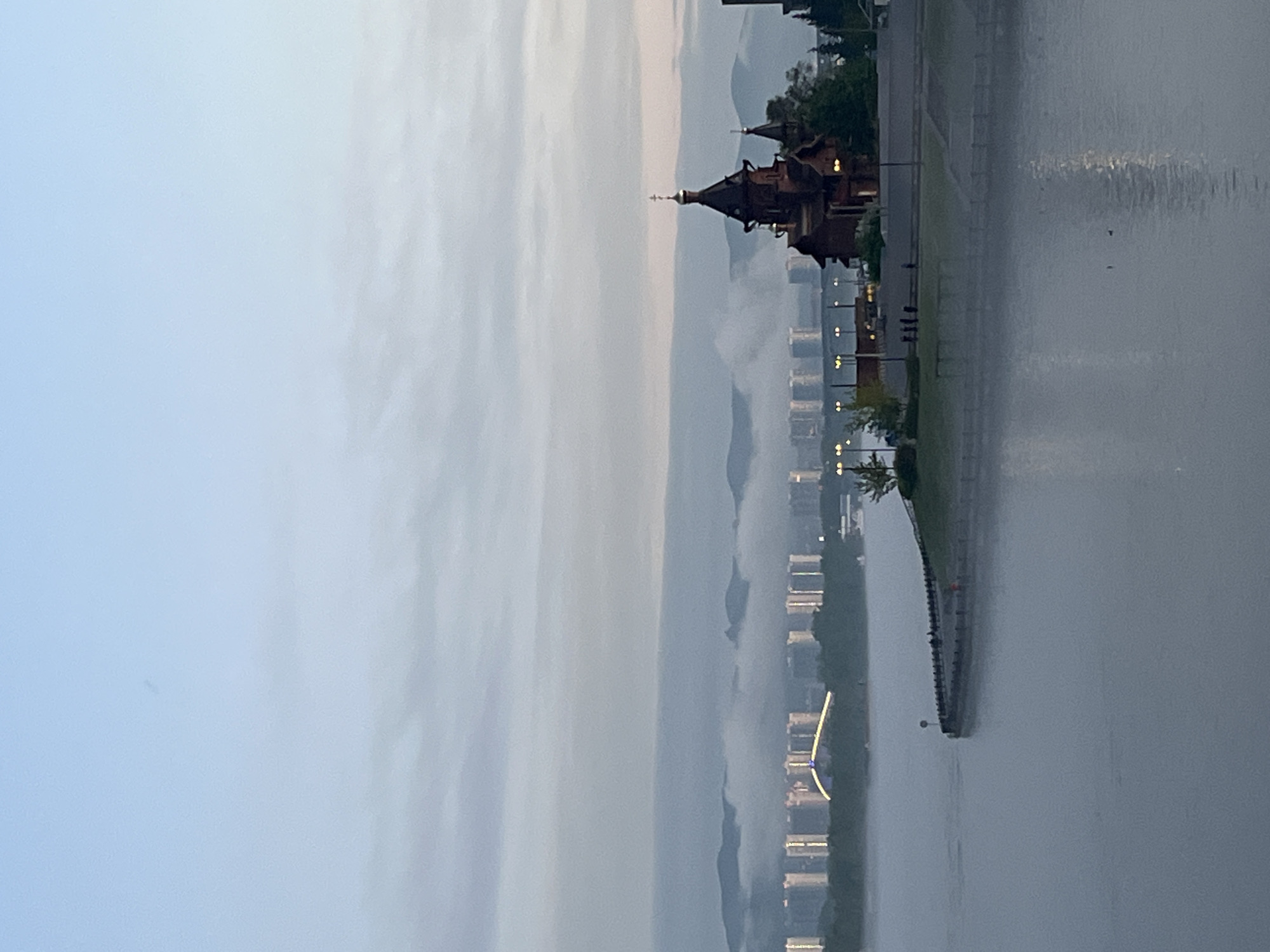 The image shows a cityscape alongside the Yenisey River in Krasnoyarsk, Russia, at dawn or dusk. The river reflects the dim light of the sky. A prominent structure resembling a traditional Russian Orthodox church or wooden palace is visible along the riverbank, possibly part of a recreational or historical complex. Several modern high-rise buildings line the river, partially obscured by fog or haze. The scene includes a paved embankment with sparse vegetation. No people are clearly visible.