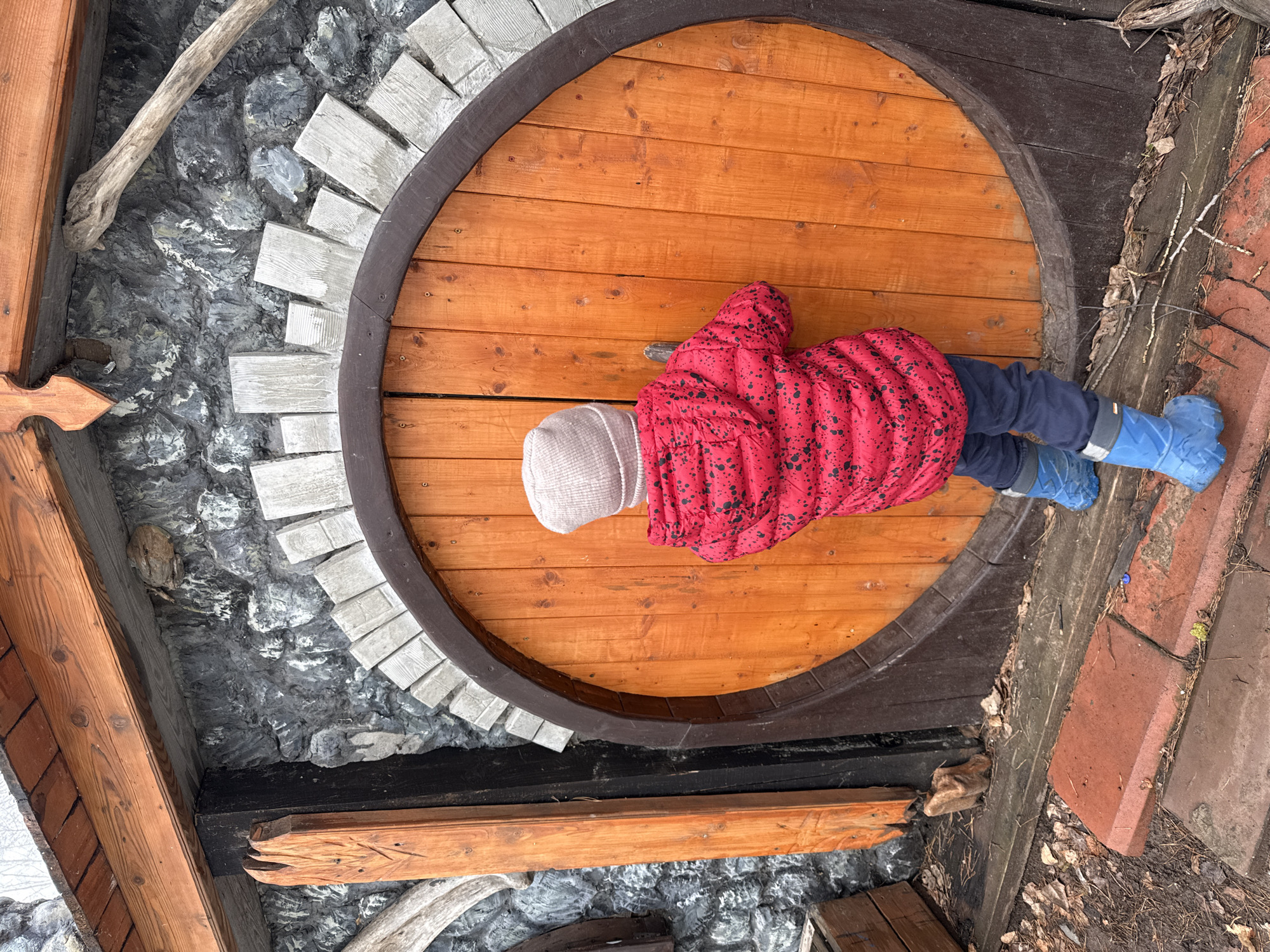 A low-angle shot depicts a child lying prone on a large, circular wooden door, possibly an entrance to a small structure, likely in the city of Chaikovsky, Russia, based on visual clues. The child is wearing a light-colored knit hat, a red puffy jacket with black spots, dark pants, and blue rubber boots. The door is made of orange wood planks and has a dark brown circular frame. The door is set within a structure featuring stone and wood elements. To the left of the door, a wall appears to be constructed of grey stone and white rectangular blocks. Wooden beams are visible above the door, suggesting an overhead structure. To the right of the door, a section of ground is visible, with what appear to be brown tiles or bricks. Dried leaves and vegetation are scattered around the ground. The lighting suggests it is daytime, but the scene appears overcast.