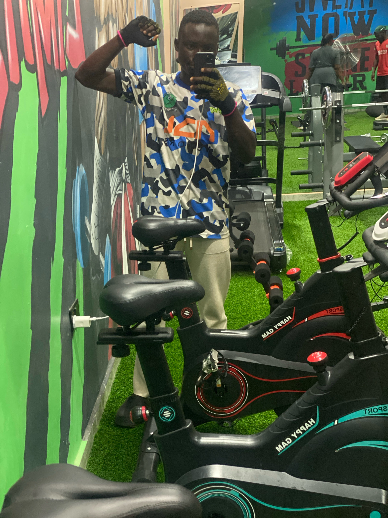 A young Black man wearing a camouflage-patterned shirt and light-colored pants is standing in a gym, taking a selfie in a mirror. He is wearing a black weightlifting glove on his right hand, with a pink band around his wrist. The gym environment includes various exercise equipment, such as stationary bikes and treadmills, with a bright green artificial turf floor. In the background, other gym-goers can be seen, and motivational text is visible on the wall. The overall scene depicts a fitness facility. The location is stated as Barden Yana, Nigeria.