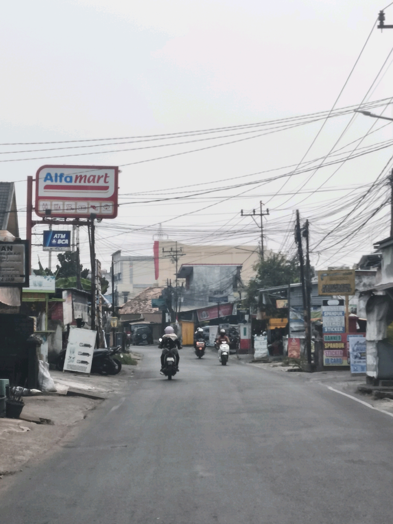 This is a slightly overcast, eye-level, medium shot of a street scene in Palembang, Indonesia. The road is paved and grey, with a white line marking the right edge in the foreground. On the left side of the road, there is a concrete sidewalk. Several motorcycles are visible on the road, some with riders. On the left, there's a large red and white sign for "Alfamart" and an ATM sign. Various buildings line the street, with some appearing to be shops or commercial establishments. Many power lines crisscross the sky above, creating a dense network. The overall atmosphere suggests a typical urban street in a Southeast Asian city.
