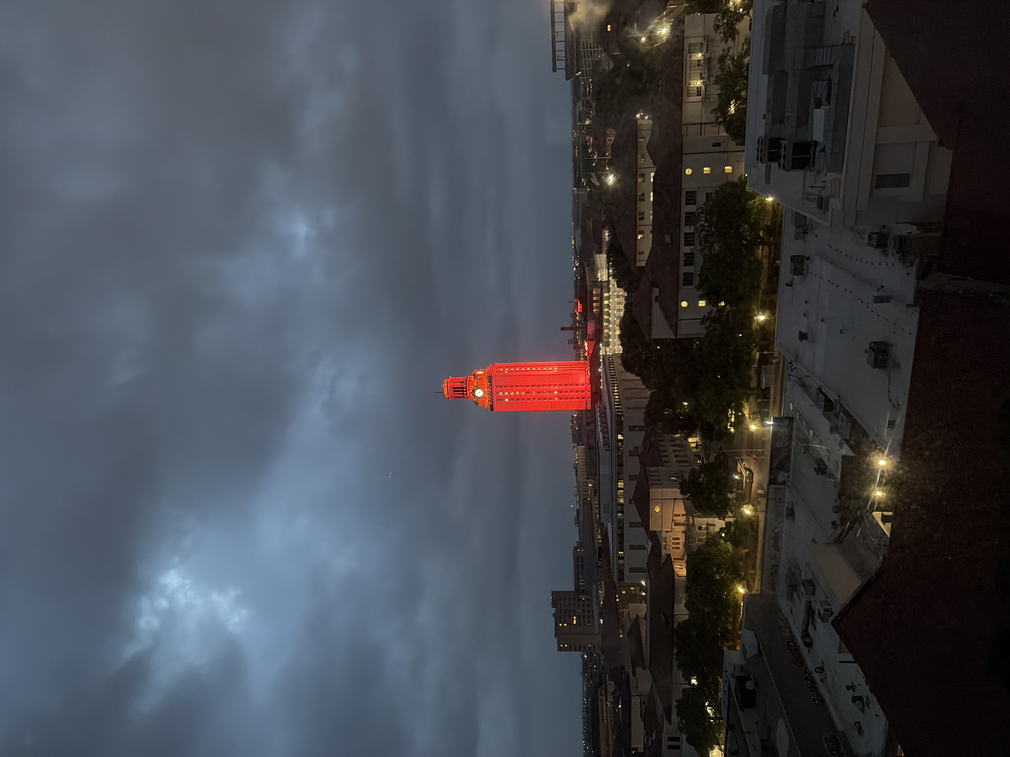 The image shows an aerial view of the city of Austin, Texas at night. The University of Texas Tower is illuminated in red and dominates the skyline. The sky is cloudy. Other buildings and streetlights are visible.