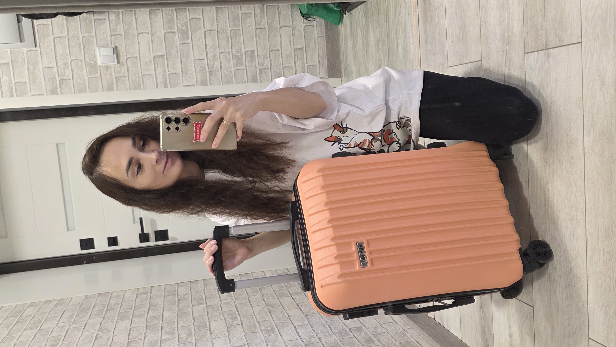 A young woman with long, dark, wavy hair is taking a selfie in a mirror. She is wearing a white t-shirt with a graphic print of cats and black pants. She is holding a peach-colored suitcase with black wheels and a retractable handle. The suitcase has a small metallic emblem with text. The woman is kneeling on a light-colored wooden floor. The background appears to be an indoor setting, possibly a hallway or a room with a brick-patterned wall on the left and a white door with black hardware on the right. A light switch is visible on the brick wall. A green bag is partially visible in the reflection on the right. The scene does not contain any elements that suggest the location is Bulgakovo, Russia.