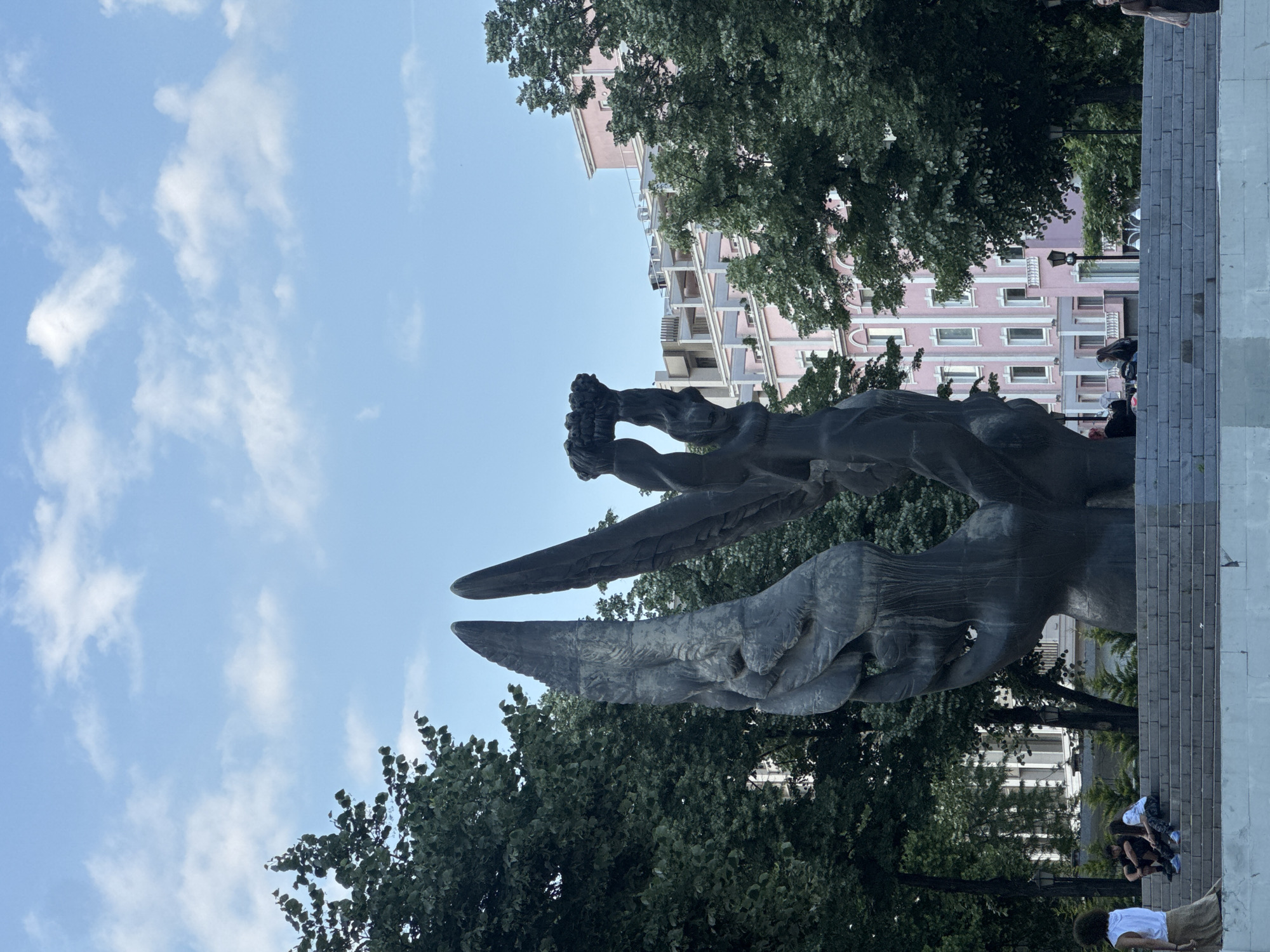 The image depicts the statue of Alyosha in Plovdiv, Bulgaria. The statue, made of dark metal, dominates the foreground, with trees surrounding it. In the background, a pink building rises behind the statue. Above, a blue sky is filled with scattered white clouds. The statue appears to have two large, stylized wings extending upwards. People sit and stand on the steps below the statue.