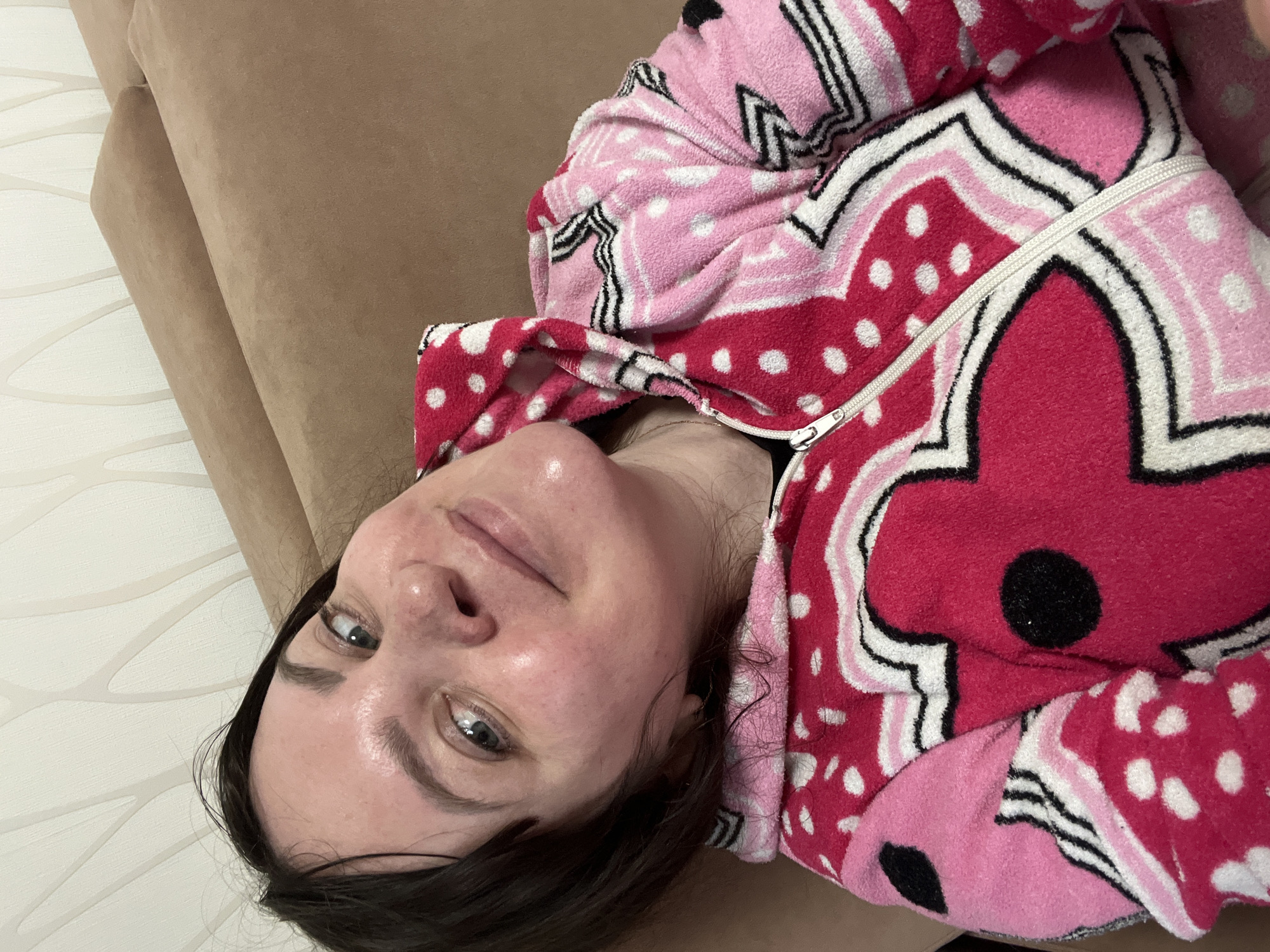 A woman lies on a brown sofa wearing a pink and red patterned bathrobe. Her dark hair is visible against the beige background of the sofa, and her face is turned towards the camera. The scene is indoors. There is no information available in this image about the location being in Perm, Russia.