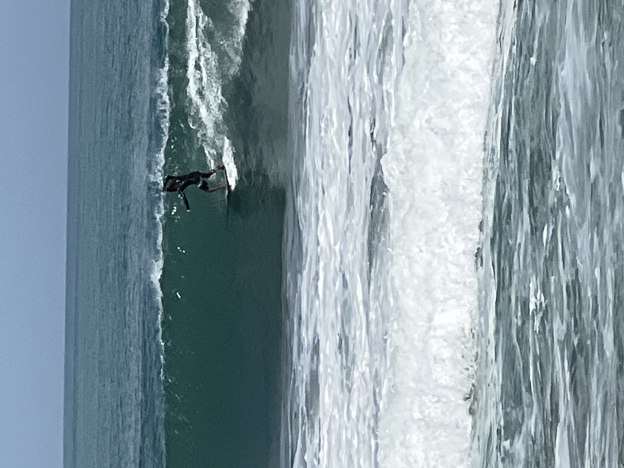 A surfer is riding a wave in the ocean. The surfer is wearing a black wetsuit and is standing on a surfboard. The wave is green and blue, with white foam cresting at the top. The sky is a pale blue. The scene appears to be a popular surfing spot, with other waves visible in the background. The location is the city of Kuta, Indonesia.