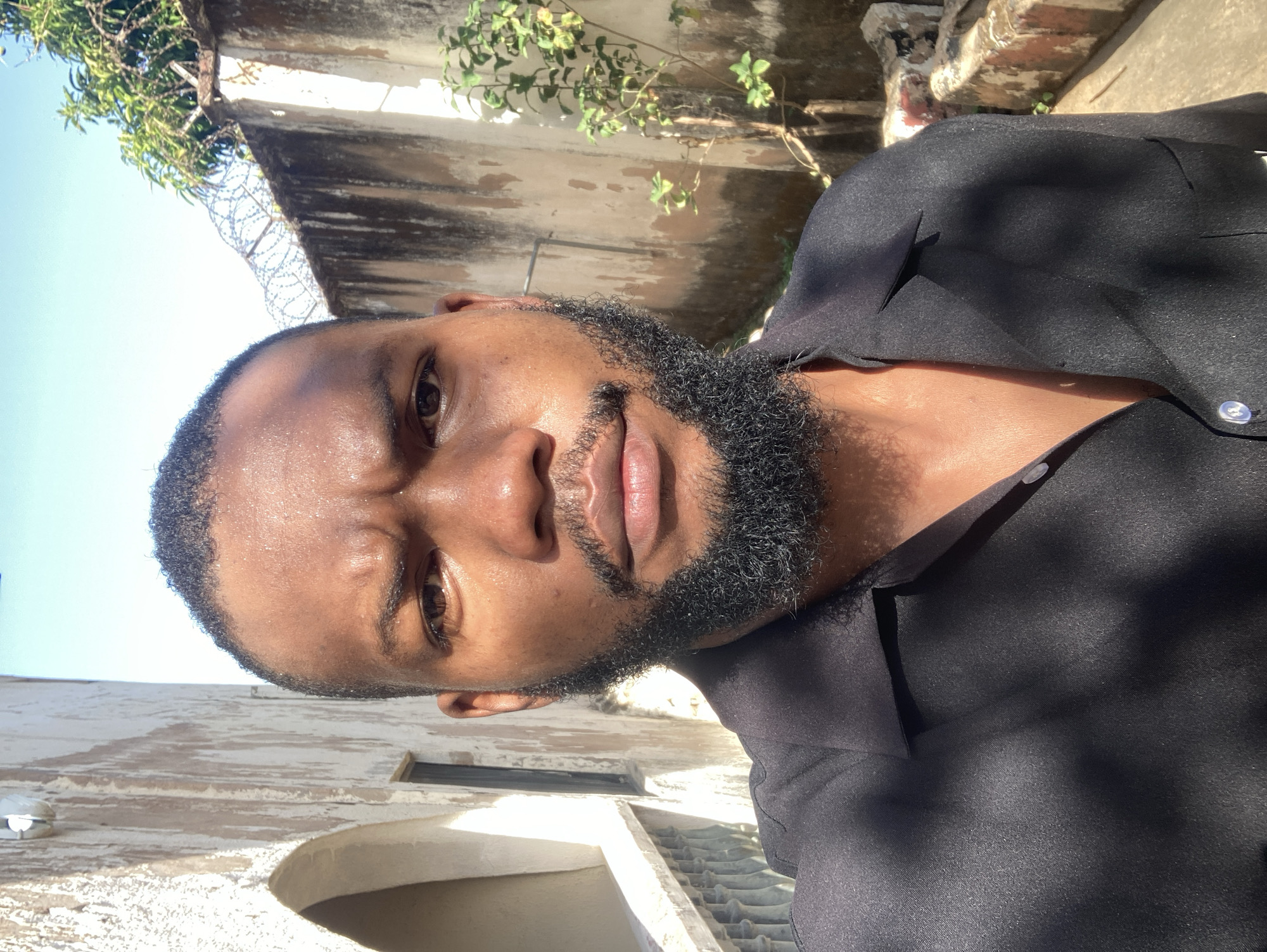A man with dark skin and a beard is shown in a medium close-up, wearing a black button-up shirt. The background suggests an outdoor location, possibly in Jos, Nigeria, based on the architectural style. A building with weathered walls, including an arched doorway and window, is visible. Razor wire is also visible on the roof of the building. The sky is light blue and appears to be sunny. The man looks directly at the camera with a serious expression.