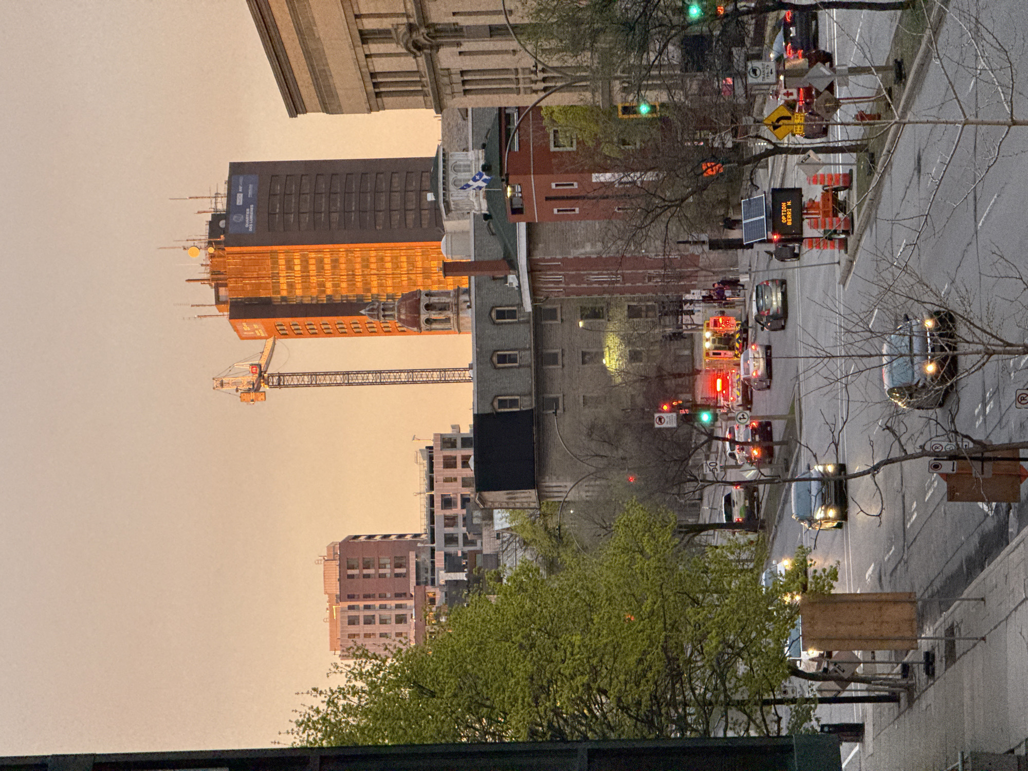 The image shows a street scene in Montreal, Canada at dusk. A major thoroughfare is visible with multiple lanes of traffic moving away from the viewer. Cars, trucks, an ambulance, and construction vehicles are present in the traffic. On the right side of the street is a sidewalk with trees and street signage. Buildings of varying heights and architectural styles line the street, including a building under construction with a crane visible. The sky is hazy, possibly due to smoke or pollution. The Quebec flag is visible atop one of the buildings. A construction sign reads "Option Berry N." There are no visible people in the street or on the sidewalks.
