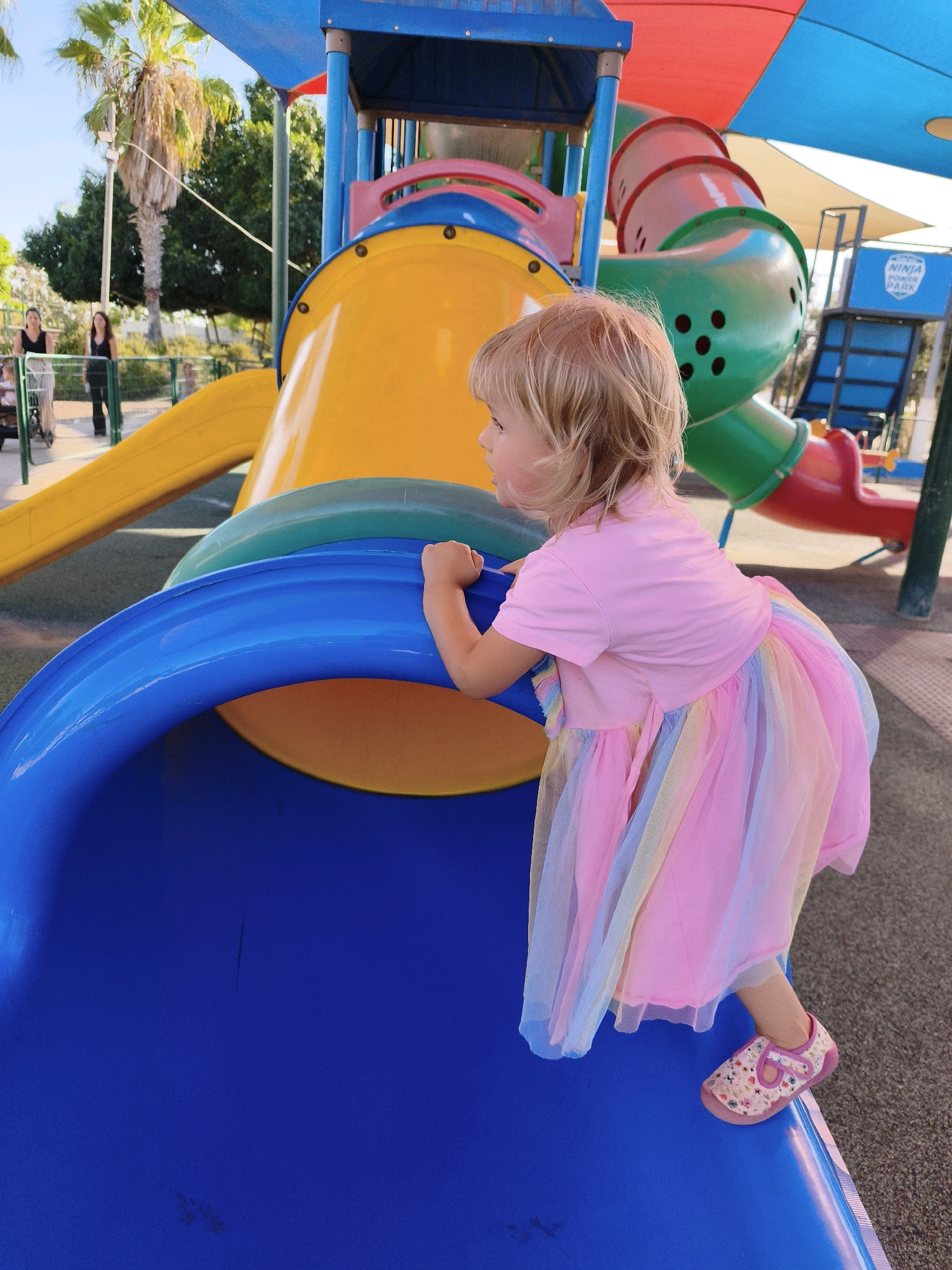 In a playground in Lachish Regional Council, Israel, a young Caucasian girl with blonde hair is on a blue and yellow slide. She is wearing a pink t-shirt and a rainbow-colored skirt. Her right hand is on the blue edge of the slide as she looks to her right. In the background, palm trees and other foliage are visible, suggesting a warm climate. The playground structure is colorful, with yellow, green, red, and blue elements. Two women, partially visible, stand in the background on the left side of the image, facing the playground. The ground of the playground is covered with a soft, gray material.