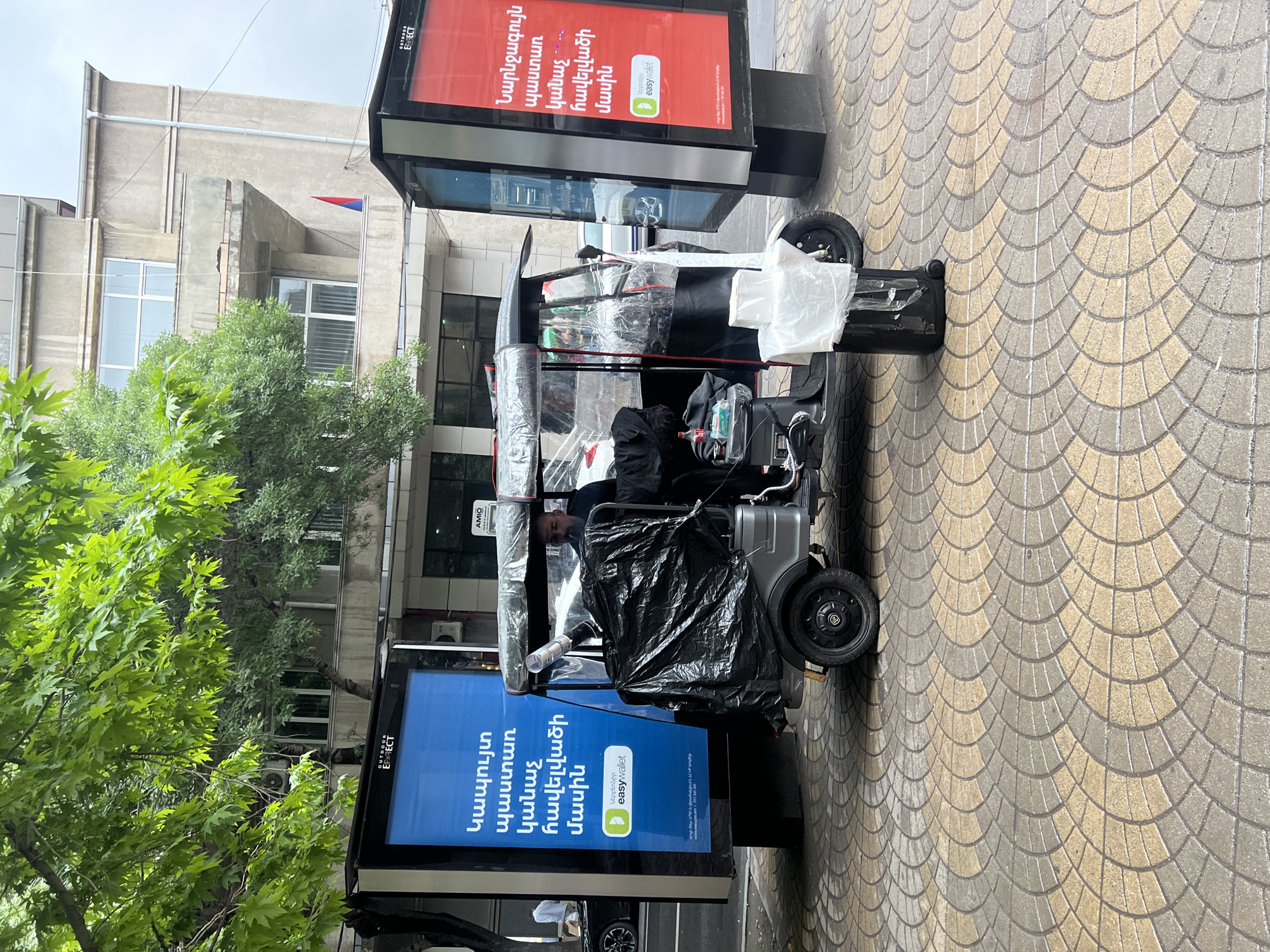 The image shows a motorized cart laden with various items, including plastic coverings and bags, positioned between two advertising displays on a city sidewalk with patterned paving stones. The advertisements feature Armenian text and logos for "easywallet," with one display having a blue background and the other having a red background. Green trees are visible to the left, along with a multi-story building in the background with windows and air conditioning units. A flag is visible on the building. Based on the street design and the presence of Armenian text, the location is likely in the city of Yerevan, Armenia.