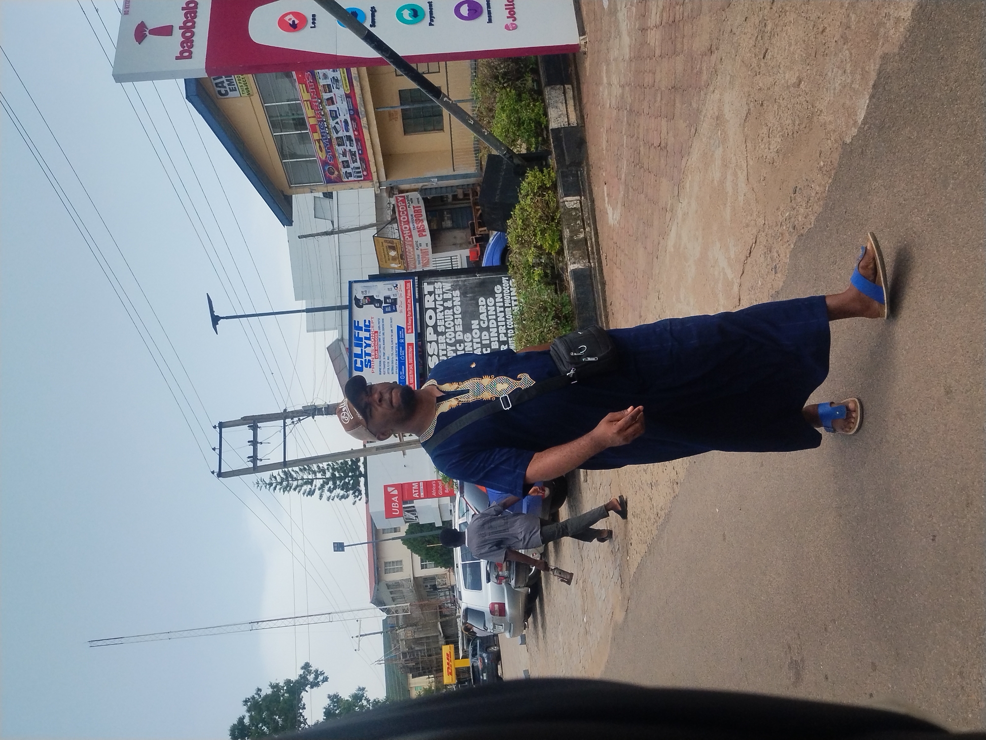 This image captures a street scene in Jos, Nigeria, with a man in traditional attire walking along the sidewalk. He is wearing a dark blue, long-sleeved robe with ornate gold embroidery on the neckline and sleeves, and a brown cap. He has a dark sling bag slung across his chest and is wearing blue sandals. The man appears to be looking towards the camera with a slight smile.
Behind him, other people are visible, including a person in a grey shirt and dark pants walking with their back to the camera, holding a brown object. In the background, there are buildings with various signs, including "baobab," "UBAN ATM Africa's Global Bank," and "CLIFF STYLIC PRINTS & CONCEPT." Other signs appear to advertise printing services like "PHOTOCOPY PHOTOCOPY PASSPORT" and "COLOR & B/W PRINTING." A white SUV and other vehicles are parked on the side of the road.
The setting suggests a commercial area, possibly a street lined with shops and businesses. The weather appears to be overcast, indicated by the bright sky and soft lighting. The overall impression is a glimpse of daily life in an urban Nigerian environment.
