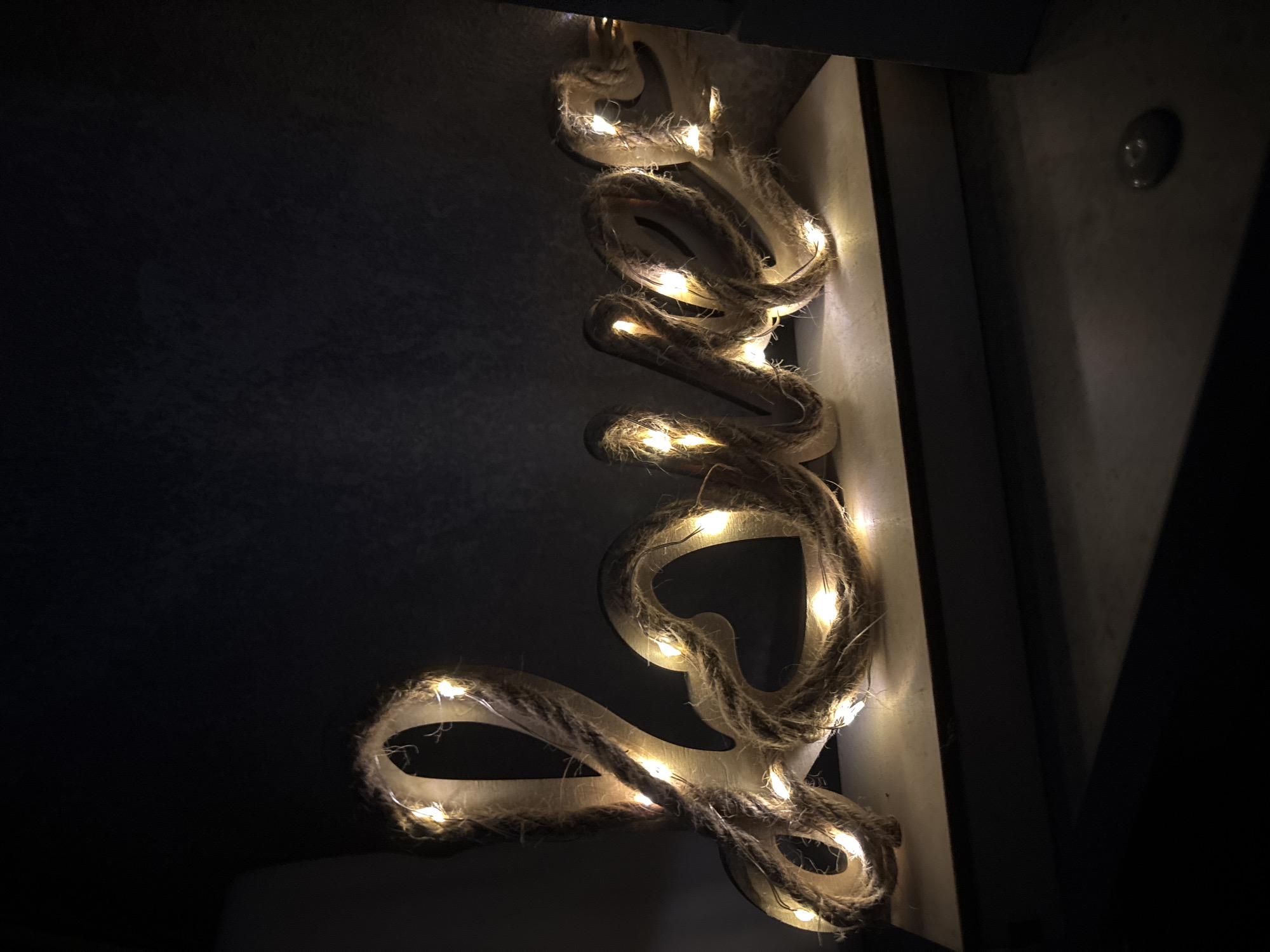 A wooden sign spelling out "Love" with rope and lights is displayed in an indoor setting. The sign is lit by a string of warm white LED lights that are woven around the rope. The letters are cut from a light-colored wood. The sign is placed on a white surface with a dark background. The overall lighting is dim, with the lights on the sign being the primary light source. The image appears to be taken in a home or a decorative display.