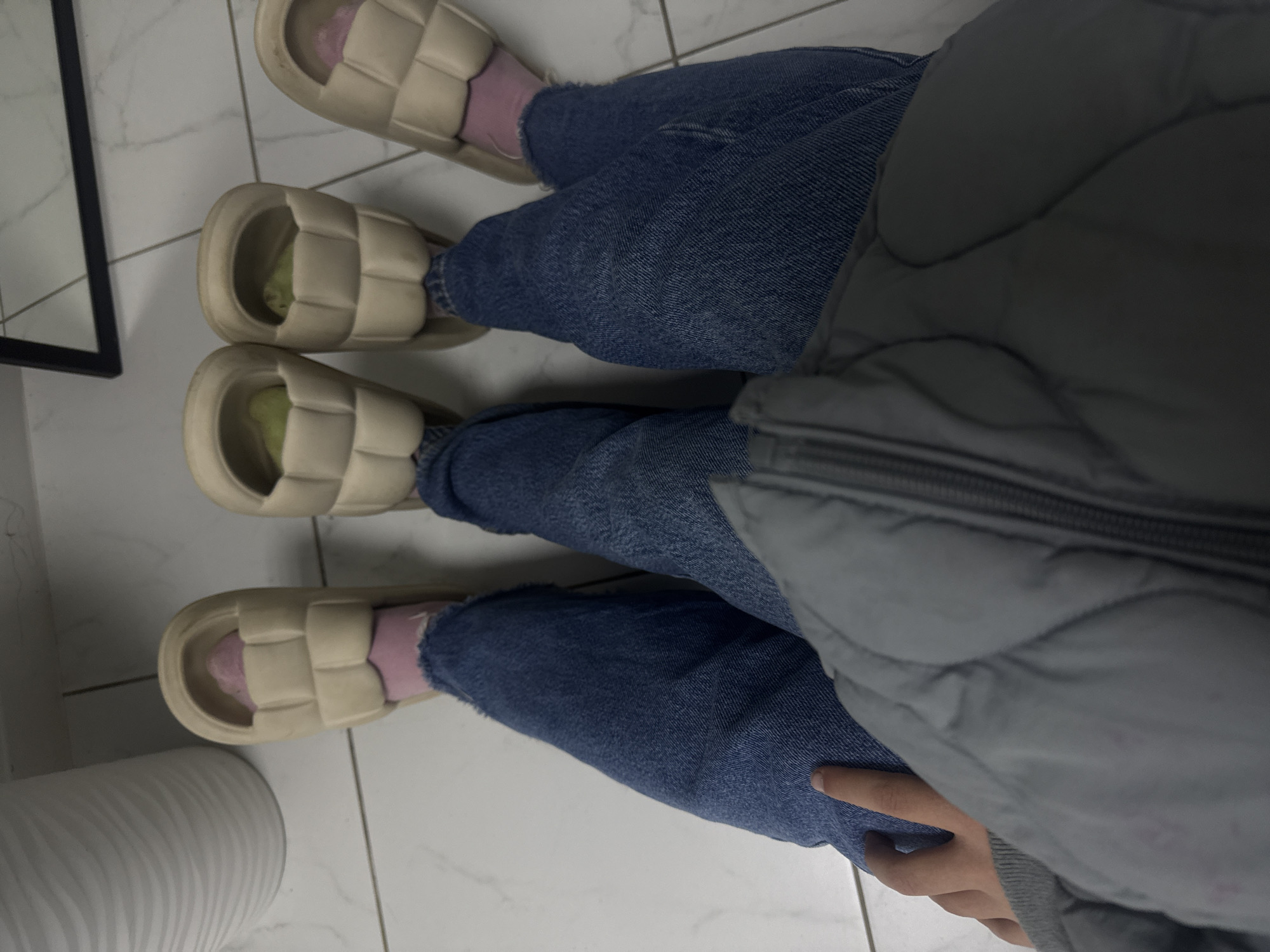 The image depicts a view from above, showing two pairs of legs wearing light blue jeans and beige, puffy sandals. One pair of legs has pink socks visible in the sandals, while the other pair appears to have lime green socks. A gray puffer jacket is partially visible, covering part of the right leg of one figure. The scene is set on a white, tiled floor. The location in the city of Votkinsk, Russia, cannot be determined from the information presented.
