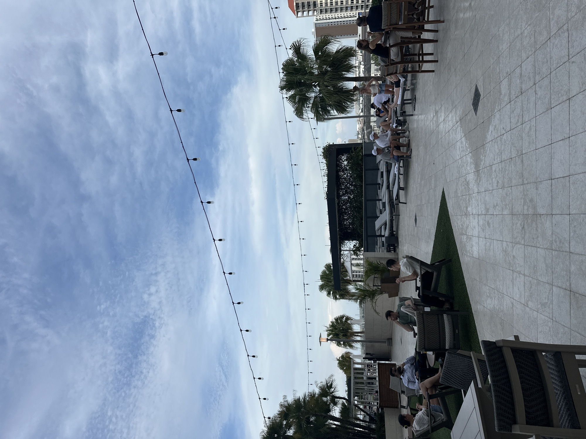 The image depicts an outdoor lounge or patio area, possibly a rooftop setting, with a view of the city skyline in Tampa, Florida, USA. The scene is captured during daylight, under a sky with scattered clouds and suspended string lights.
People are seen sitting on various chairs and benches, either alone or in small groups. They are engaged in leisurely activities like conversation, relaxation, or using electronic devices. The patio features a combination of tiled flooring and patches of artificial turf. Potted plants and palm trees add to the ambiance.
The scene includes architectural elements such as pergolas or shaded structures, which provide a break from the sun. In the background, taller buildings of the city are visible, giving context to the location. The presence of outdoor furniture and the general setting indicates a commercial or recreational space for gatherings.