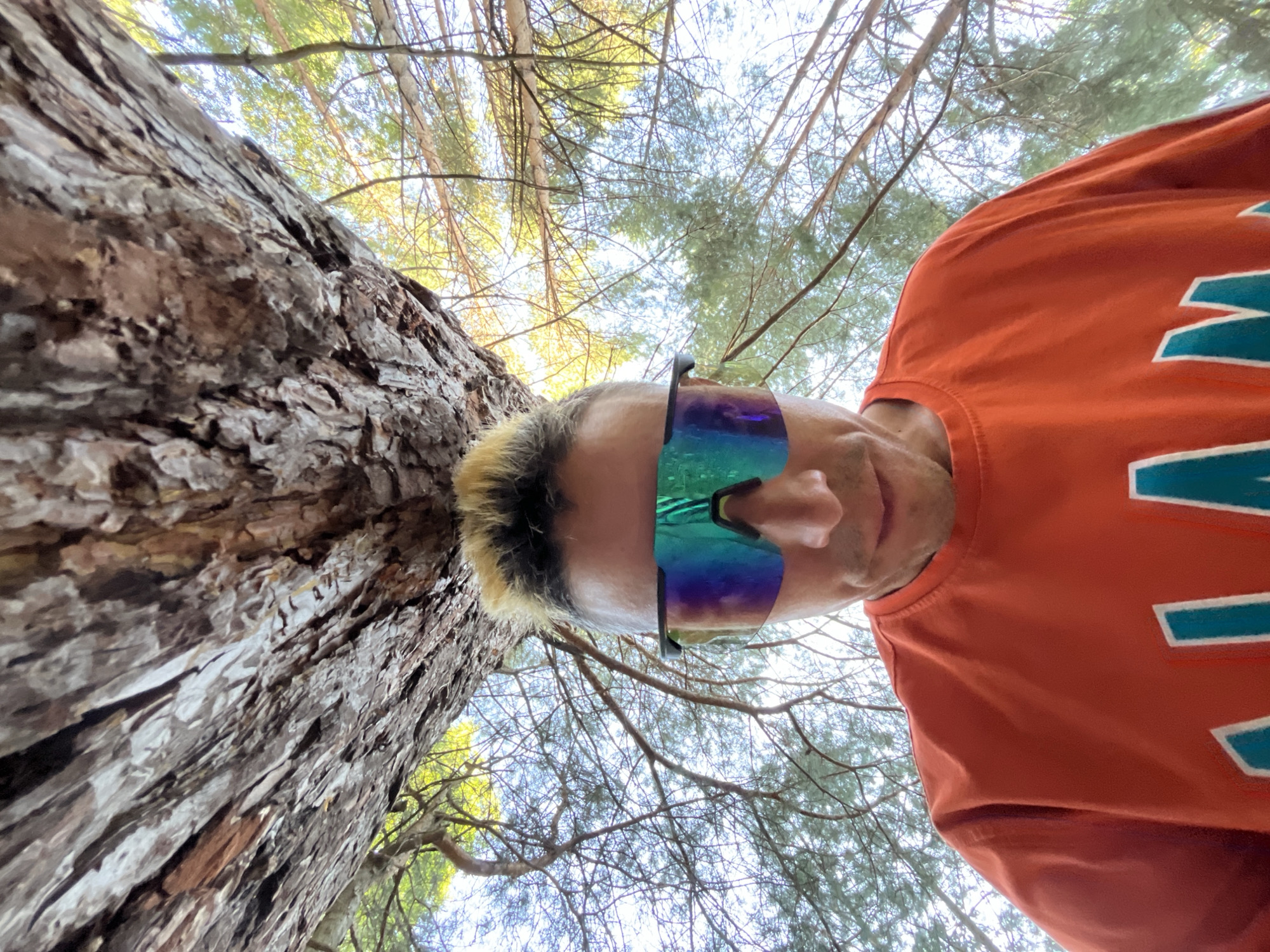 A low-angle, close-up, downward-facing shot shows a person's face and torso in an outdoor setting, likely a forest given the presence of a large tree trunk and branches overhead. The person is wearing mirrored wrap-around sunglasses that reflect the sky and trees. They are also wearing an orange t-shirt with a teal and white graphic design. The image is a selfie taken from a unique perspective, looking up at the subject from below. There is no indication of the location being in the city of Chaikovsky, Russia.