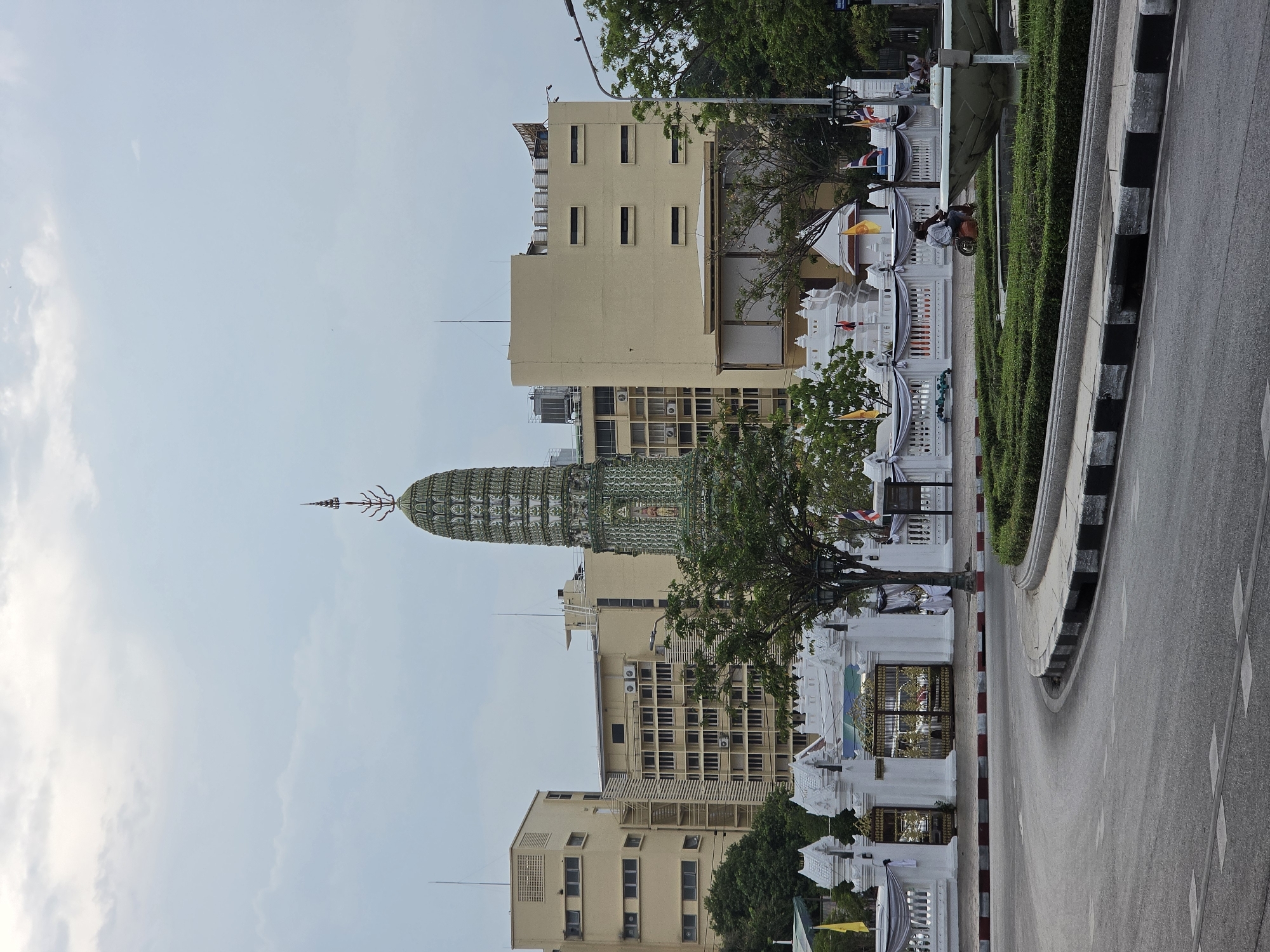 The image shows a view of the temple of Wat Ratchabophit, with its distinctive emerald green and white pagoda, in Bangkok, Thailand. The temple is surrounded by cream-colored buildings and a tree, while a white fence runs along the sidewalk. A road is visible on the right side of the image. The sky is light blue with some clouds. A moped is parked next to the sidewalk.