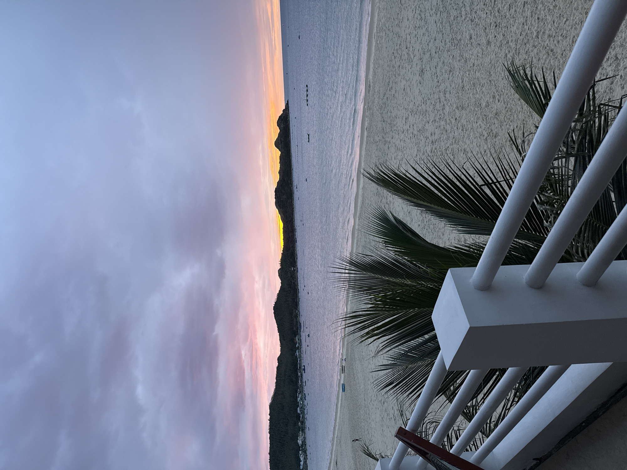 This is a wide-angle, eye-level shot of a beach scene at dusk in Song Cau, Vietnam. In the foreground, a white railing with a square post runs across the bottom of the frame, obscuring some of a sandy beach and palm fronds. Beyond the railing, the sand stretches to the water's edge. The sea is calm and a muted blue, with several small boats scattered across its surface. In the distance, a line of dark, tree-covered hills forms the horizon, separating the sea from the sky. The sky is filled with soft, diffused clouds painted in shades of pale pink, lavender, and grey, with a band of warm orange and yellow light near the horizon, suggesting the sun has recently set. The overall impression is one of tranquility and natural beauty.