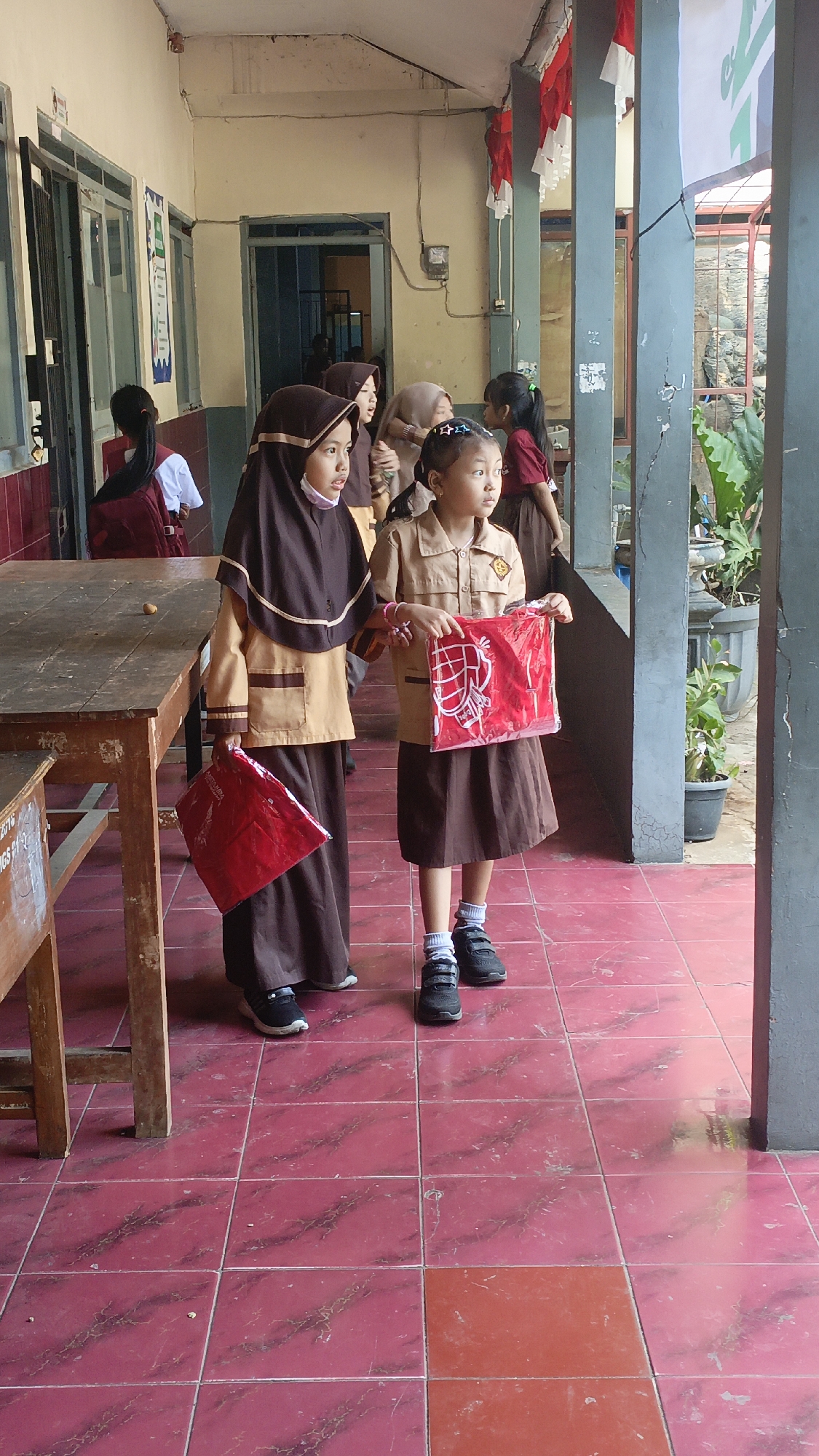 Two young girls in school uniforms are standing on a tiled floor under a covered walkway. The girl on the left wears a brown hijab and a beige and brown uniform. She holds a red bag in her left hand. The girl on the right wears a beige and brown uniform and holds a red bag in front of her. Both girls are looking to their right. Behind them, other students in school uniforms can be seen. The walkway has large pillars, and red and white flags are hanging from the ceiling, suggesting a national celebration. The floor is tiled in red with a marble-like pattern. The environment appears to be a school in Salatiga, Indonesia, given the context provided.