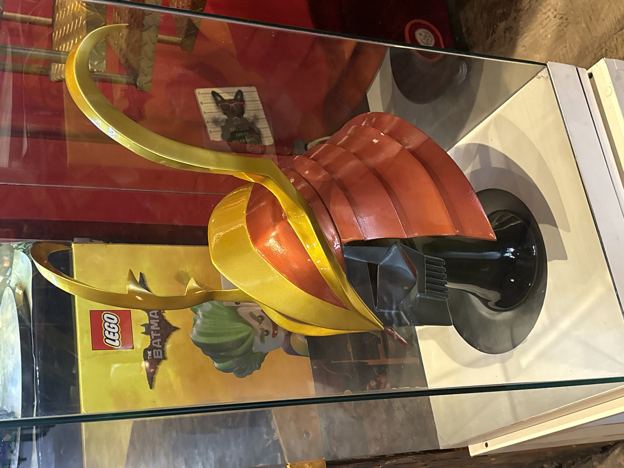 The image shows a display case in what appears to be a museum or a themed shop. Inside the glass case, a replica of a helmet, likely a samurai helmet, is prominently displayed on a black stand. The helmet is an elaborate design with a black base, a red lacquered section that resembles layered plates, and a gold-colored crest with sweeping curves.
Behind the helmet, a yellow box with a red LEGO logo is visible. Below the LEGO logo, a silhouette of the Batman symbol is printed, suggesting the helmet is related to Batman merchandise or a Batman-themed exhibit. A Lego version of the Joker with green hair and a mischievous grin can be seen on the box.
The background consists of red walls and shelves. Another display with what looks like a framed picture or artwork featuring a black silhouette against a wood-grain background is visible on the right side of the case. The display case itself is made of glass and sits on a white platform.
Based on the provided context, this display is located at the "Museu Pelé" (Pelé Museum) in Santos, São Paulo, Brazil. This specific exhibit is likely part of a collection related to "The Batman" franchise, possibly incorporating elements from the Lego Batman movie or other related merchandise, as indicated by the presence of the Lego box and the Batman symbol. The museum, while primarily dedicated to the legendary footballer Pelé, occasionally hosts temporary exhibitions of various themes.