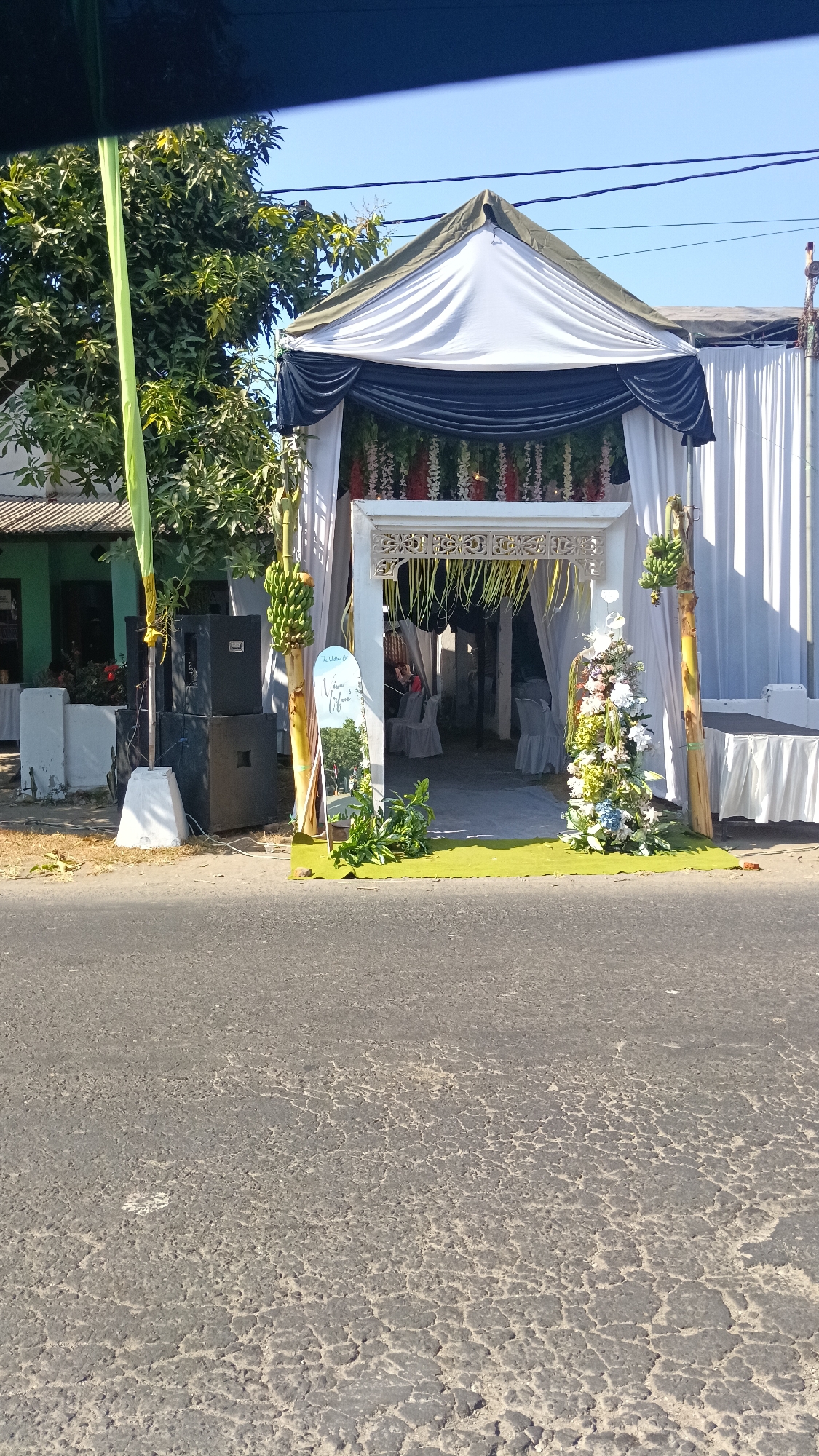 The image depicts an outdoor wedding setup in front of a building, likely a house, in the city of Dukuh, Indonesia. The foreground shows a paved road. In the middle ground, a white tent with dark blue drapes is erected, serving as the main event space. The entrance to the tent is decorated with hanging flowers and greenery. To the left of the entrance, there are large black speakers stacked on a concrete base and a banana tree with a bunch of bananas. A decorative sign with text "The Wedding of Vera & Fifaar" stands near the entrance, supported by a banana tree. To the right of the entrance, another banana tree is present, and further to the right, white drapes cover the side of a building and a white-covered table. Inside the tent, chairs are set up, indicating a seating arrangement for guests. The background shows trees, the building facade, and a clear blue sky. The overall scene suggests a festive occasion, possibly a wedding reception, taking place outdoors.