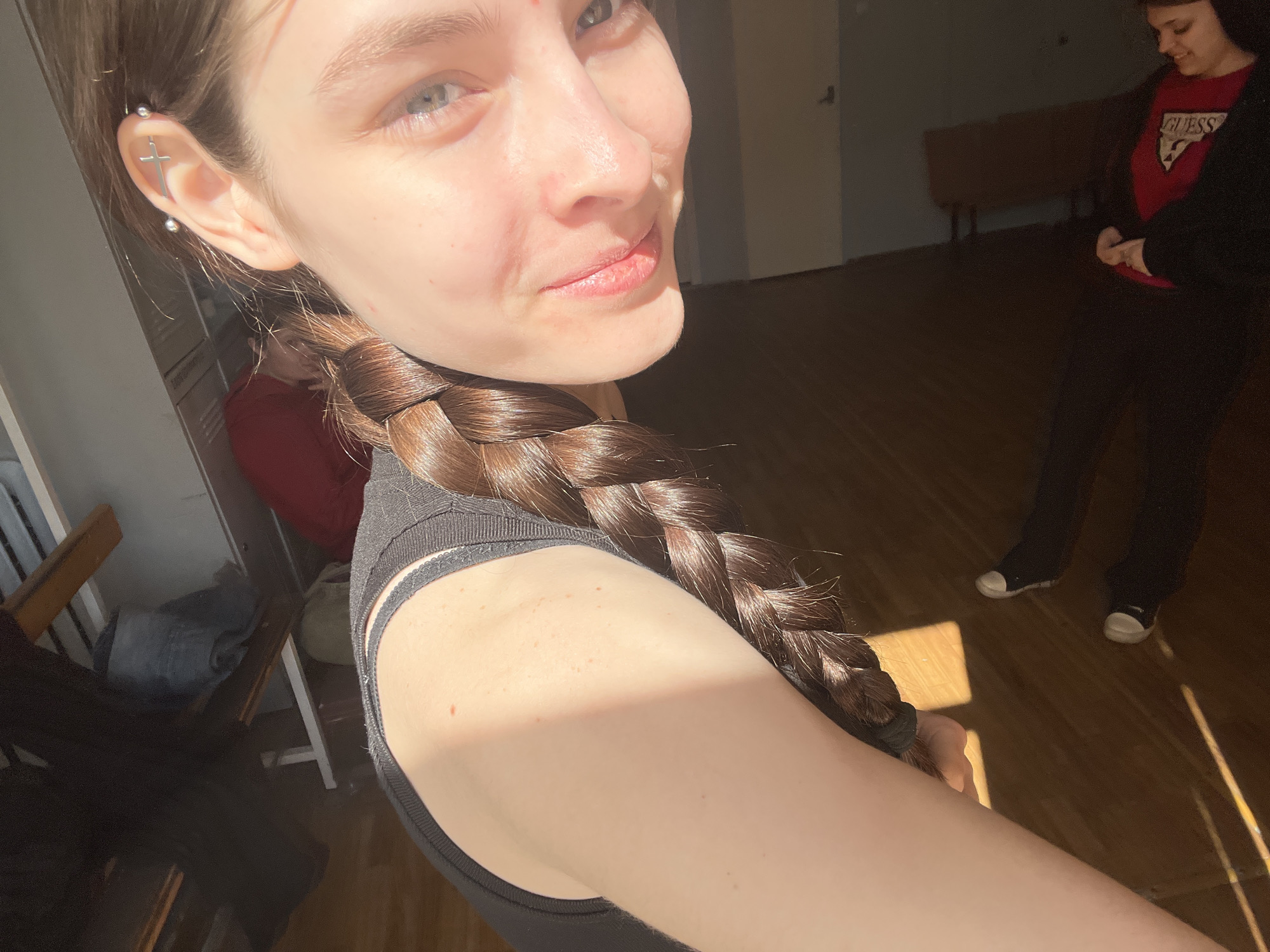 The image captures a close-up selfie of a young woman, illuminated by strong natural sunlight entering from the left. She is the primary subject, shown from the chest up, looking directly at the camera with a subtle, serene smile. Her light skin is visible, and her blue eyes are bright. Her long, dark brown hair is styled in a thick braid, draped over her left shoulder and chest. A notable detail is a silver industrial piercing in her left ear, shaped like a cross. She wears a black, ribbed sleeveless top or sports bra.
In the background, another person is partially visible on the right, standing and looking downwards. This individual wears a red t-shirt with the word "GUESS" printed prominently on it, paired with dark pants and sneakers. The setting appears to be indoors, likely a spacious room with warm-toned wooden floors, where sunlight casts distinct shadows, suggesting it is daytime, possibly late morning or early afternoon. Dimly seen furniture, including chairs and a bench, can be discerned in the deeper background. The overall atmosphere is relaxed. The location context provided is Saint Petersburg, Russia.