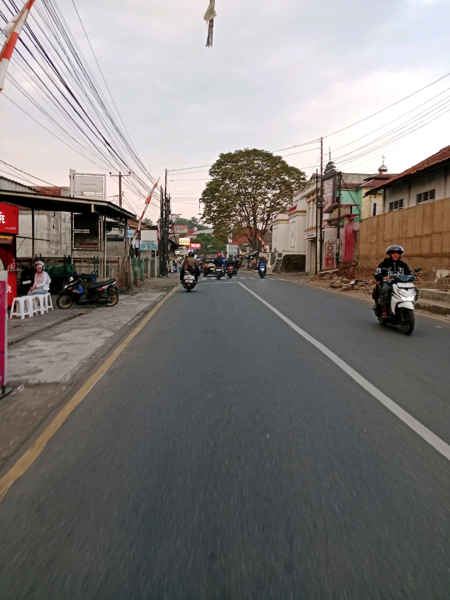 The image captures a street scene in Sirnagalih, Indonesia, likely during the late afternoon or early evening given the soft lighting. The perspective is from a moving vehicle, looking forward down a paved road lined with buildings and overhead electrical wires.
On the left side of the road, there is a building with a dark roof and a white facade. In front of it, several white plastic stools are arranged, and a person in a white head covering and white top is seated on one of them. A black scooter is parked nearby. Further down the street on the left, there is a red sign.
The road itself is asphalt with a yellow line marking the edge on the left and a white line dividing the lanes on the right. Several motorcycles are visible on the road. In the foreground, a white scooter with a rider wearing a helmet and black jacket is approaching from the right. Further ahead, multiple other scooters and motorcycles are moving in the same direction.
On the right side of the road, buildings with various colored facades, including green and beige, are visible. One building has a dome on its roof, suggesting a religious structure. A tall tree with dense foliage stands in the middle distance.
The sky is overcast, indicating a cloudy day. The overall impression is of a typical urban street with active traffic.