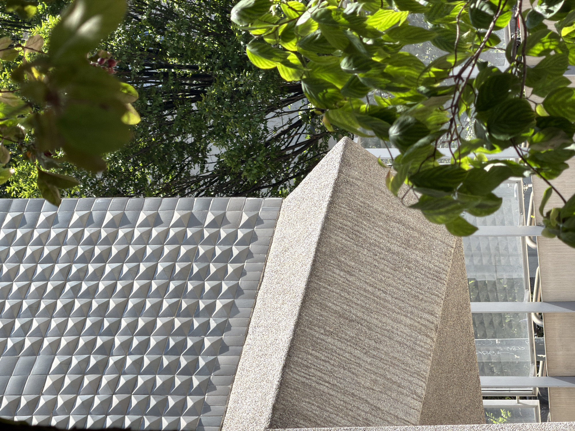 The image shows a building in Milan, Italy. The building has a textured facade with triangular protrusions on the left. Adjacent to this is a tan-colored section, textured with horizontal lines and small specks. The building appears modern in style. Overhanging the building are tree branches with green leaves, creating a natural frame. To the right of the tan section, there is a glass and metal structure, potentially a part of the building.