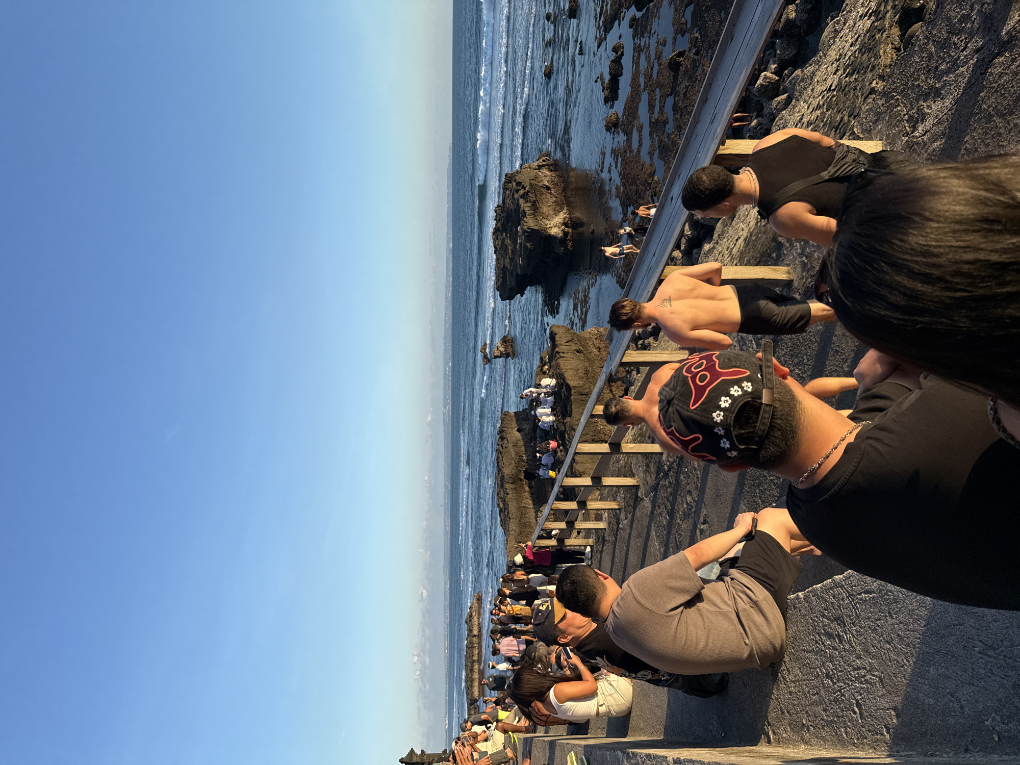 The image depicts a coastal scene in North Kuta, Indonesia, showcasing a gathering of people near the ocean. A crowd of people can be seen on the coast of North Kuta, Indonesia, near the sea. A wooden walkway is present, with some individuals walking down it towards the water. Others are observing the scene from a higher vantage point. The ocean has waves and several rocks jutting out. The sky is clear and blue.