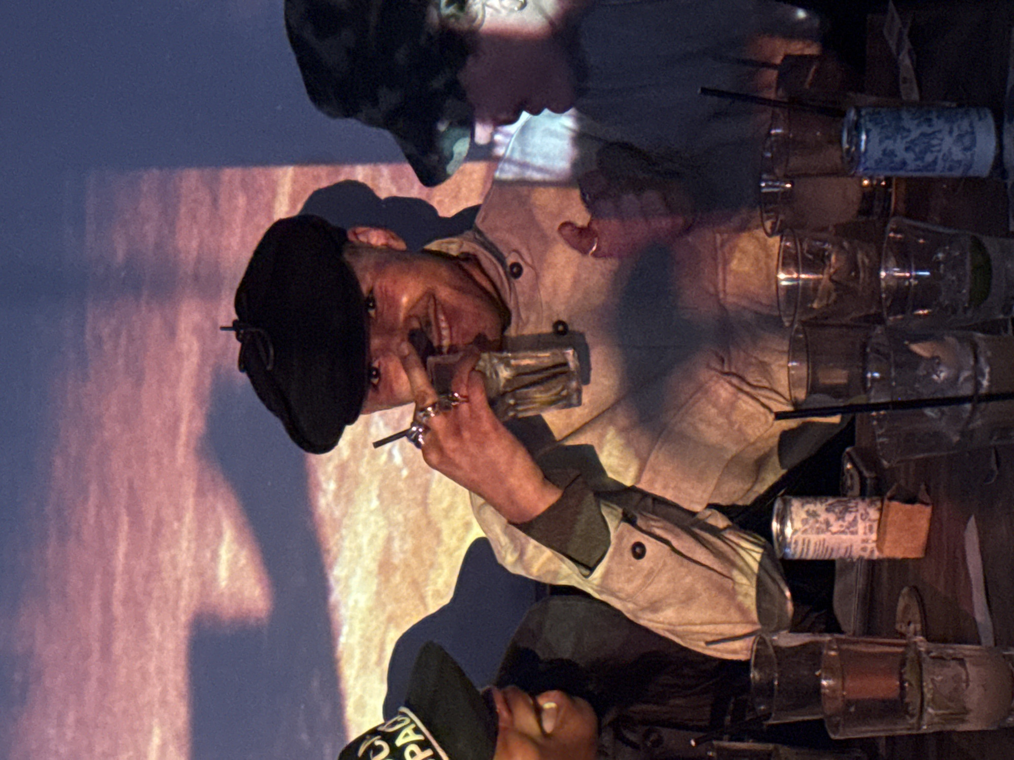 A photo depicts a man indoors, wearing a black beret and a light-colored jacket, holding a small glass container and smiling slightly. Another person in a cap is partially visible in the lower part of the frame. A variety of empty glass containers, some with straws, are aligned on what appears to be a bar or table, suggesting a social environment. The background includes blurred colors suggesting an urban environment, possibly the city of London, United Kingdom, known for its vibrant nightlife.
