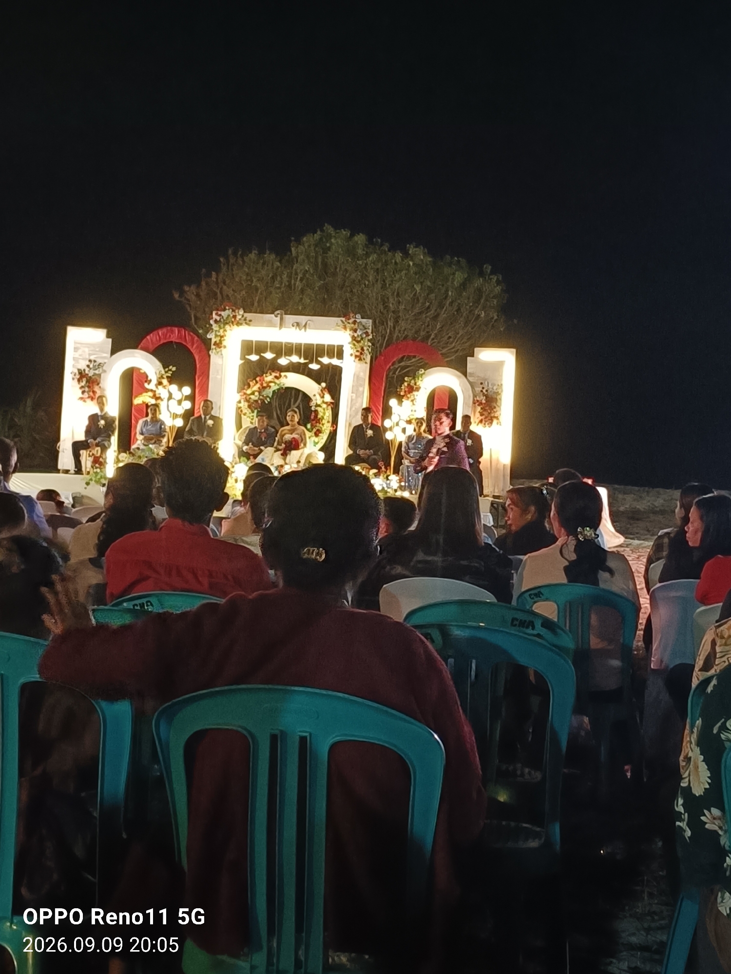 The image depicts an outdoor evening event, likely a wedding ceremony, taking place in Otan, Indonesia. In the background, a lit stage is set up with a floral arch and illuminated large letter decorations. Several individuals, possibly members of the wedding party, are seated on the stage, with a couple at the center appearing to be the bride and groom. To the left, a person is speaking into a microphone. In the foreground, a crowd of people are seated in teal-colored plastic chairs, facing the stage. The overall atmosphere appears festive, illuminated by the stage lights and the surrounding darkness. The image also includes a timestamp and device information in the bottom left corner: "OPPO Reno11 5G 2026.09.09 20:05".