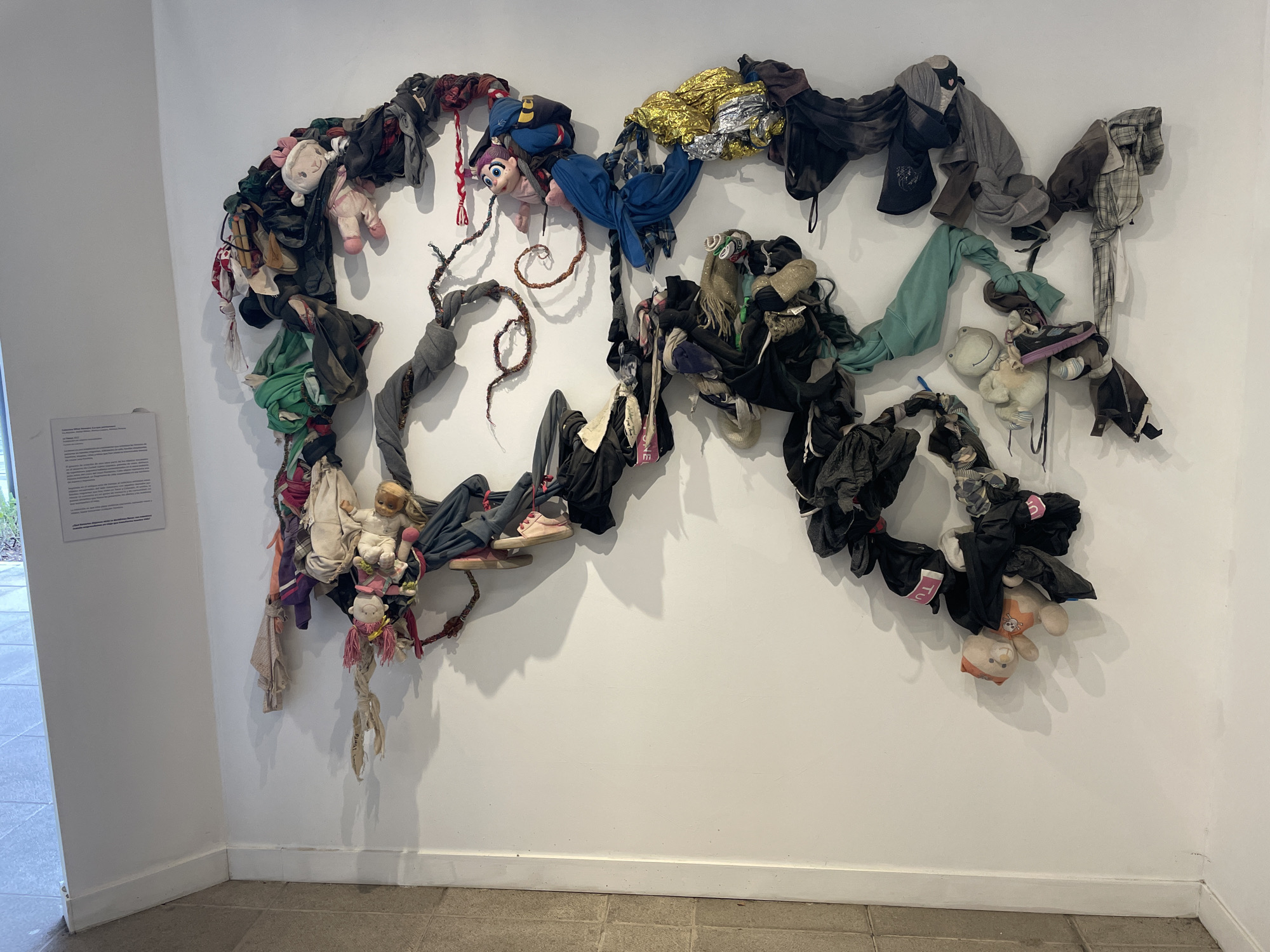 The image shows an art installation on a white wall inside a gallery in Mexico City, Mexico. The installation consists of intertwined fabric scraps, clothing items, and stuffed animals creating an abstract, wave-like shape. The scraps are a mixture of colors and textures. There is a sign attached to the wall on the left of the installation. The floor is tiled.