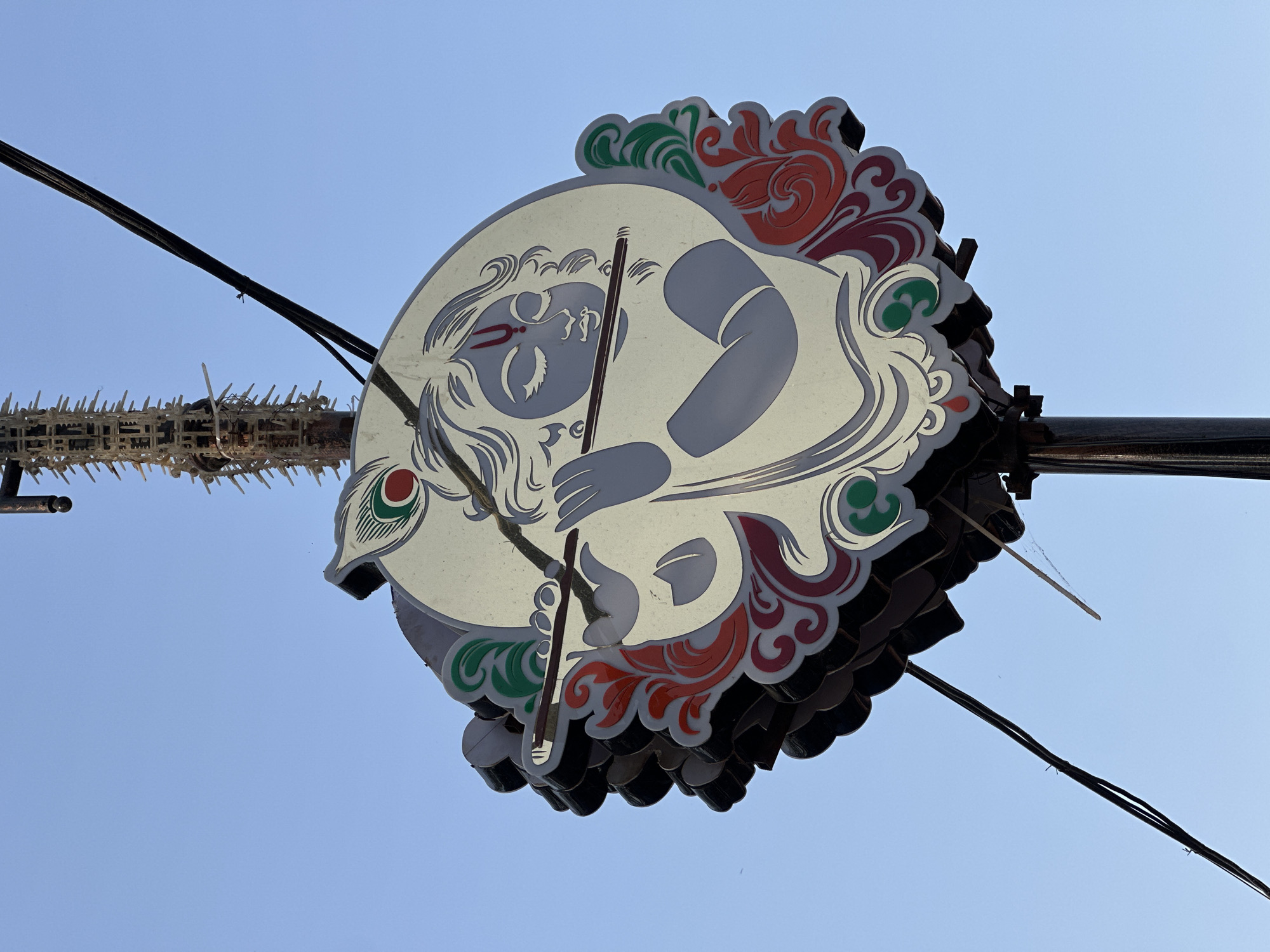 A low-angle shot shows a sign depicting Krishna in Mathura, India. The circular sign, viewed from below, features a light-colored background with a grey image of Krishna holding a flute. Red and green floral designs border the image. The sign appears to be made of multiple layers. Black electrical wires run across the blue sky in the background. A structure with pointed decorations sits near the left edge of the sign.