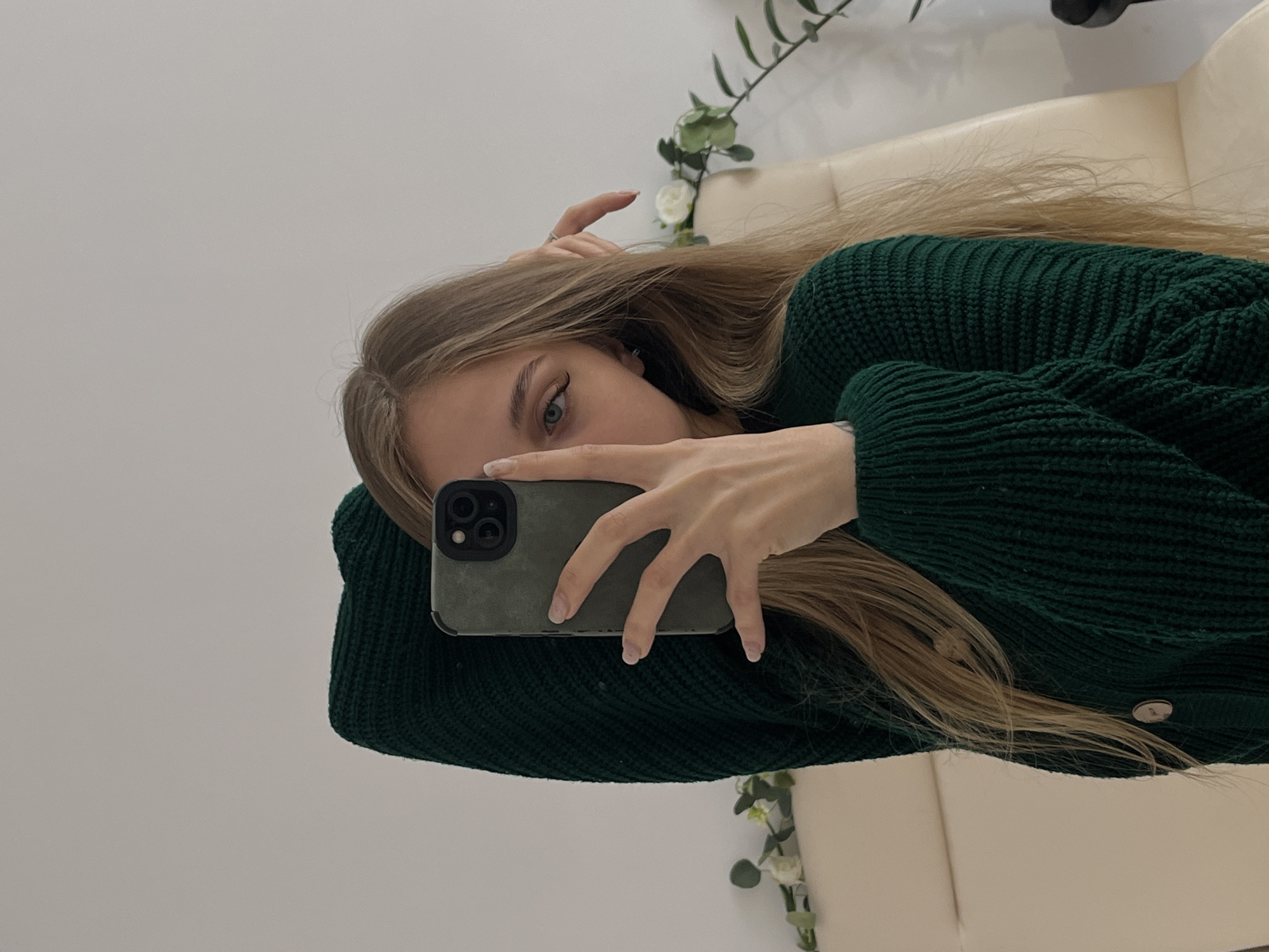 A woman is taking a selfie with a phone. She is wearing a dark green sweater. Her hair is long and blonde. A beige couch and some greenery are visible in the background. It's impossible to verify the location in Фоки, Russia from this image alone.