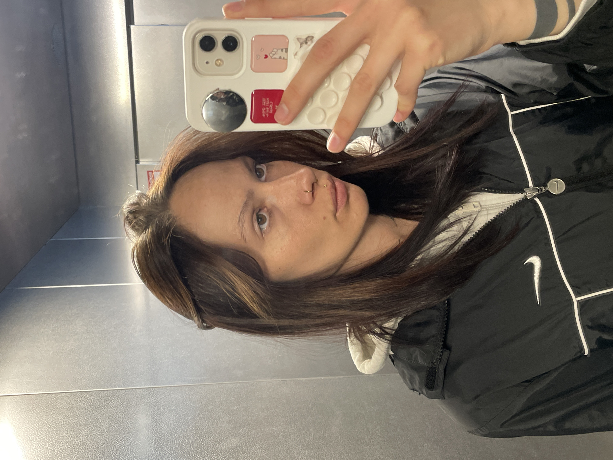 A person is taking a selfie in what appears to be an elevator. The background is a gray, tiled wall. The person is holding a white iPhone in their right hand, which partially obscures their face. They have brown hair with blonde highlights, brown eyes, and a piercing in their nostril. They are wearing a black Nike jacket with white accents over a white hoodie. Part of a wrist tattoo is visible on their left wrist.
There is no information in the image to suggest this is taken in the city of Izhevsk, Russia.