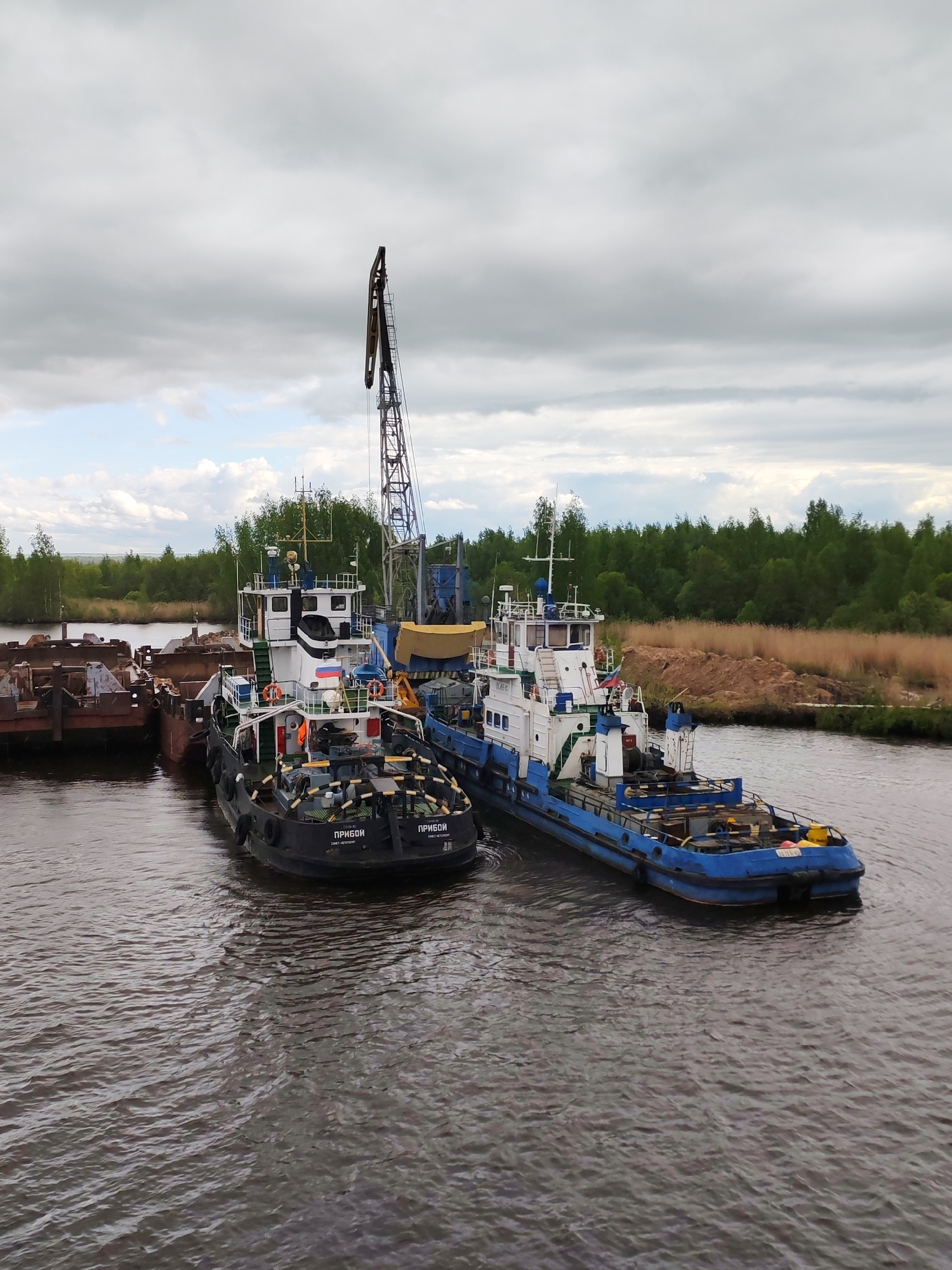 The image shows two tugboats moored in a river or waterway in the city of Ozernoe Ust'e, Russia. The tugboats, one painted black and white and the other blue and white, are docked alongside a barge or pier on the left side of the frame. A crane is visible extending upwards from one of the boats. The water is murky and dark. The background features a treeline and a cloudy sky.