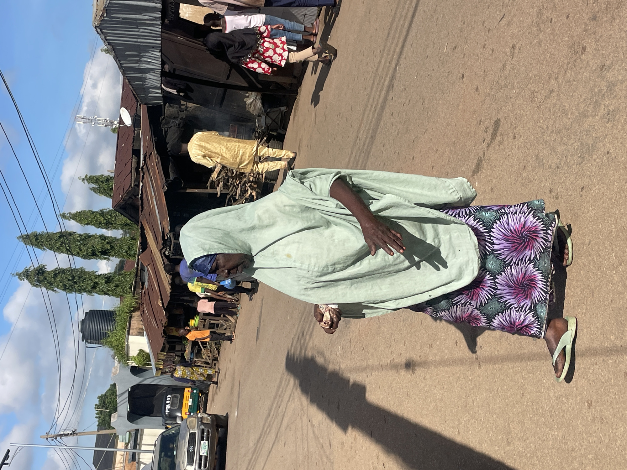 The image depicts a street scene in Jos, Nigeria, under a bright, partly cloudy sky. In the foreground, a woman wearing a light green hijab and a long, vibrantly patterned skirt walks from left to right. Her right hand is held up, appearing to hold a small object, and her left hand is near her waist. She is wearing light green flip-flops. The ground is a dusty, asphalt road.
Behind the woman, several other people are visible. To the left, a silver SUV is parked. Further back, a yellow and green motorized tricycle (Keke Napep) is partially visible. A series of market stalls with corrugated metal roofs and wooden structures line the street on the right side of the road. Men and women are seen near these stalls, some appearing to be vendors or shoppers. In the background, tall trees and a water tank are visible. A telecommunications tower with a satellite dish stands prominently in the distance, suggesting an urban or semi-urban setting. The overall impression is of a busy, informal market area on a sunny day.
