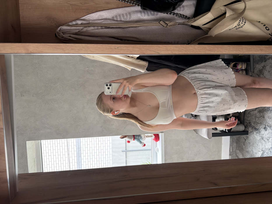 A young adult female is depicted in a mirror selfie, holding a light-colored smartphone with her left hand to capture the image. She has blonde hair styled in a ponytail, wears a white sports bra or bralette, light-colored patterned shorts with a drawstring, white wireless earbuds in both ears, and a necklace with a small cross pendant.
The background, reflected in the mirror, shows an indoor setting with light gray textured walls and a patterned tile floor. To the left, a window reveals an exterior white brick wall and a decorative figure with a red hat and pants on the windowsill. Below the window, a gray-cushioned bench or shoe rack is present, with multiple pairs of shoes visible beneath it. To the right of the mirror's reflection, inside a wooden wardrobe or cabinet, various articles of clothing, a black shoulder bag with a chain strap, and a beige canvas tote bag are visible.
The specific location within the city of Rossiyskiy, Russia, is not discernible from the visual evidence provided.