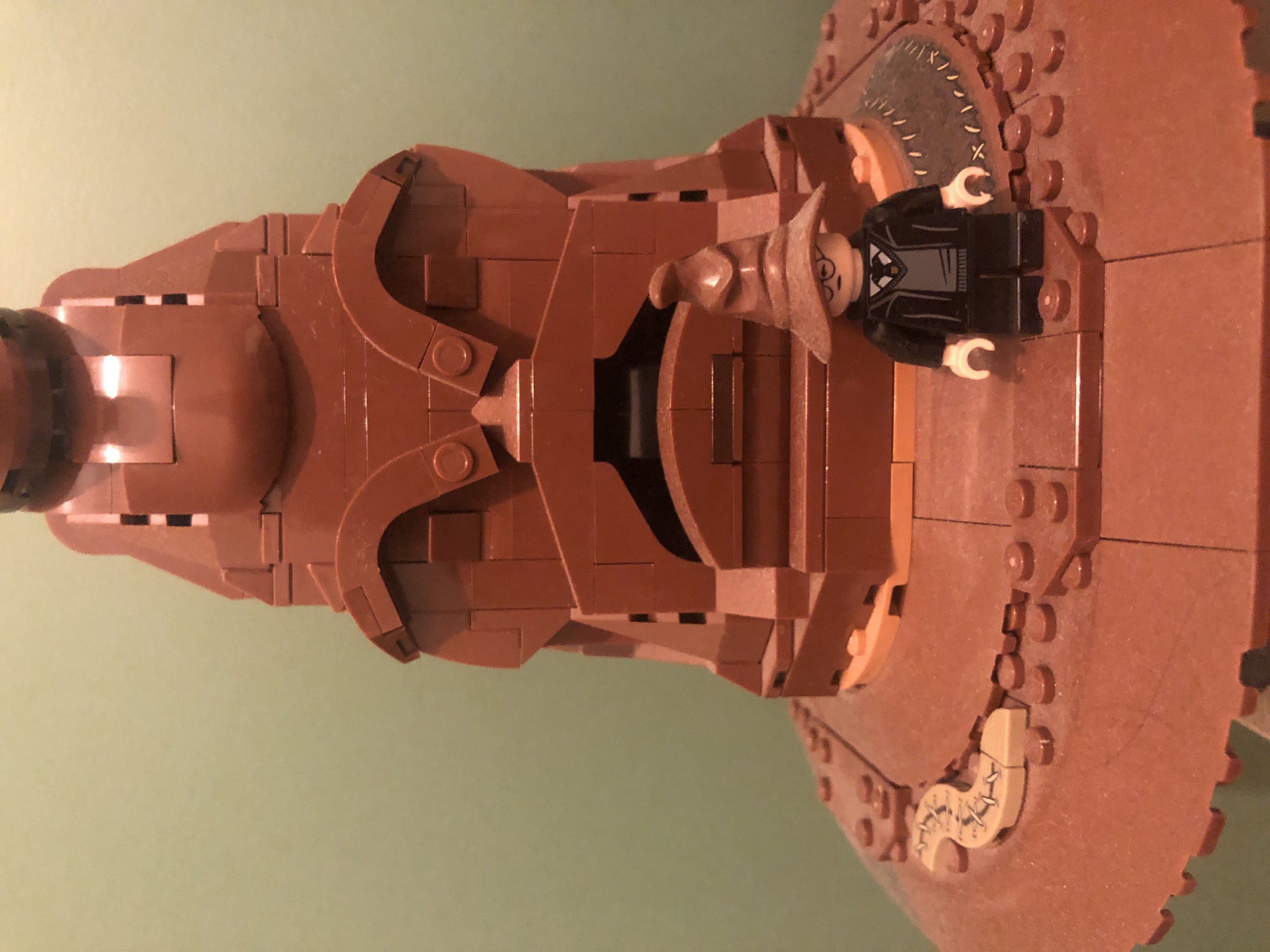 A close-up, eye-level shot shows a Lego figure of Harry Potter standing in front of a large Lego replica of the Sorting Hat. The Sorting Hat is predominantly brown, with visible brick lines and a textured surface suggesting a conical shape with facial features. The Harry Potter figure is also brown and is wearing a school uniform with a tie and glasses. The background is a solid, light green wall. The overall scene suggests a display of Lego creations.