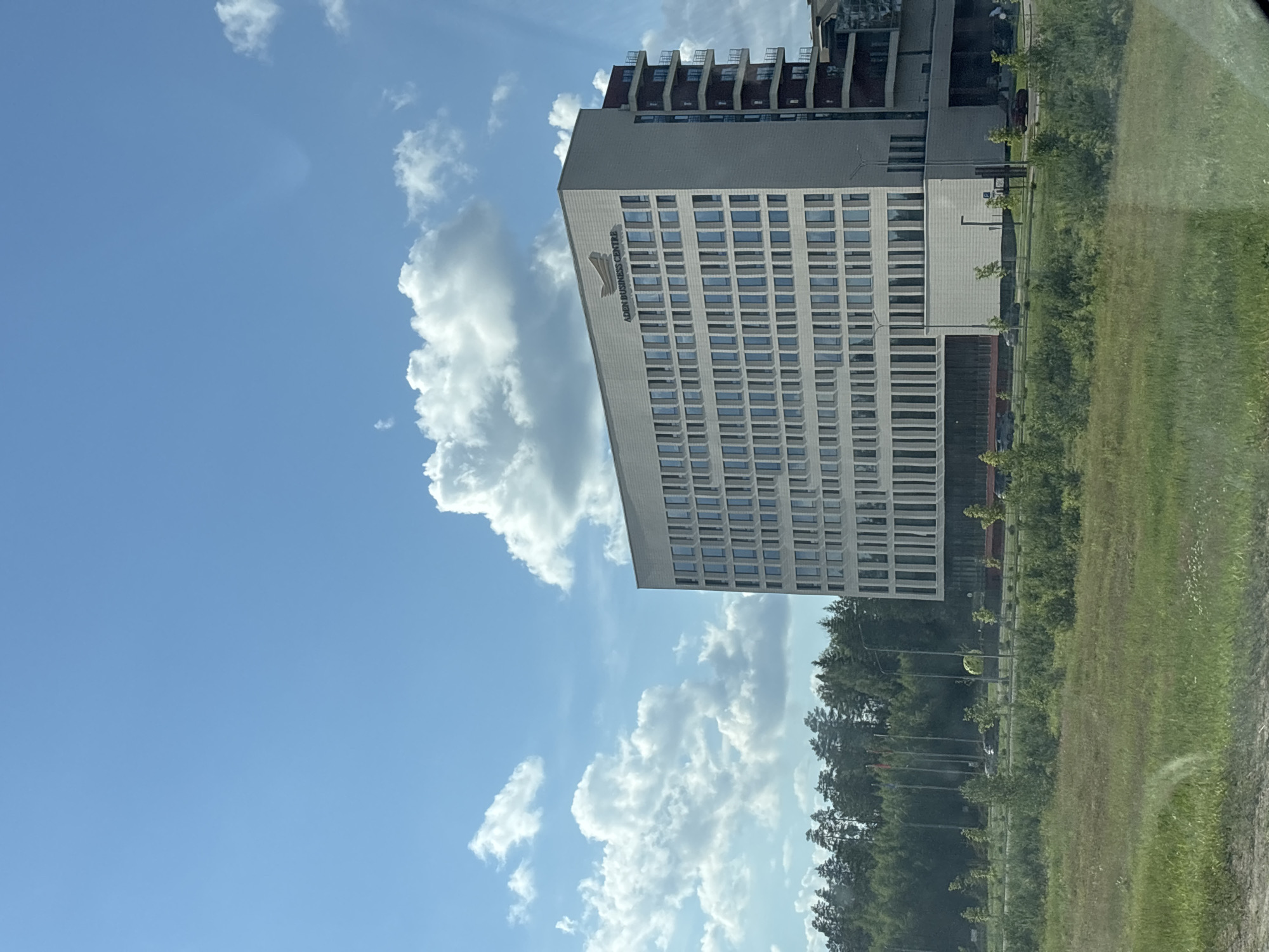 The image displays a modern, light-colored building with numerous windows, identified as the ADDI BUSINESS CENTRE by text on its facade. The building appears to be multi-story, with a darker structure visible on top. It is situated in a green, grassy area with a line of trees in the background. The sky above is a clear blue, dotted with fluffy white clouds. The scene suggests a daytime setting, possibly late morning or afternoon, with bright, natural light. There are no people visible, but there are hints of infrastructure like streetlights and potentially parking areas. The overall impression is one of an urban or semi-urban environment with modern architecture integrated with natural landscaping. The orientation of the image is landscape, but the content appears to be rotated 90 degrees counter-clockwise. The location is specified as Драчкаўскі сельскі Савет, Belarus.